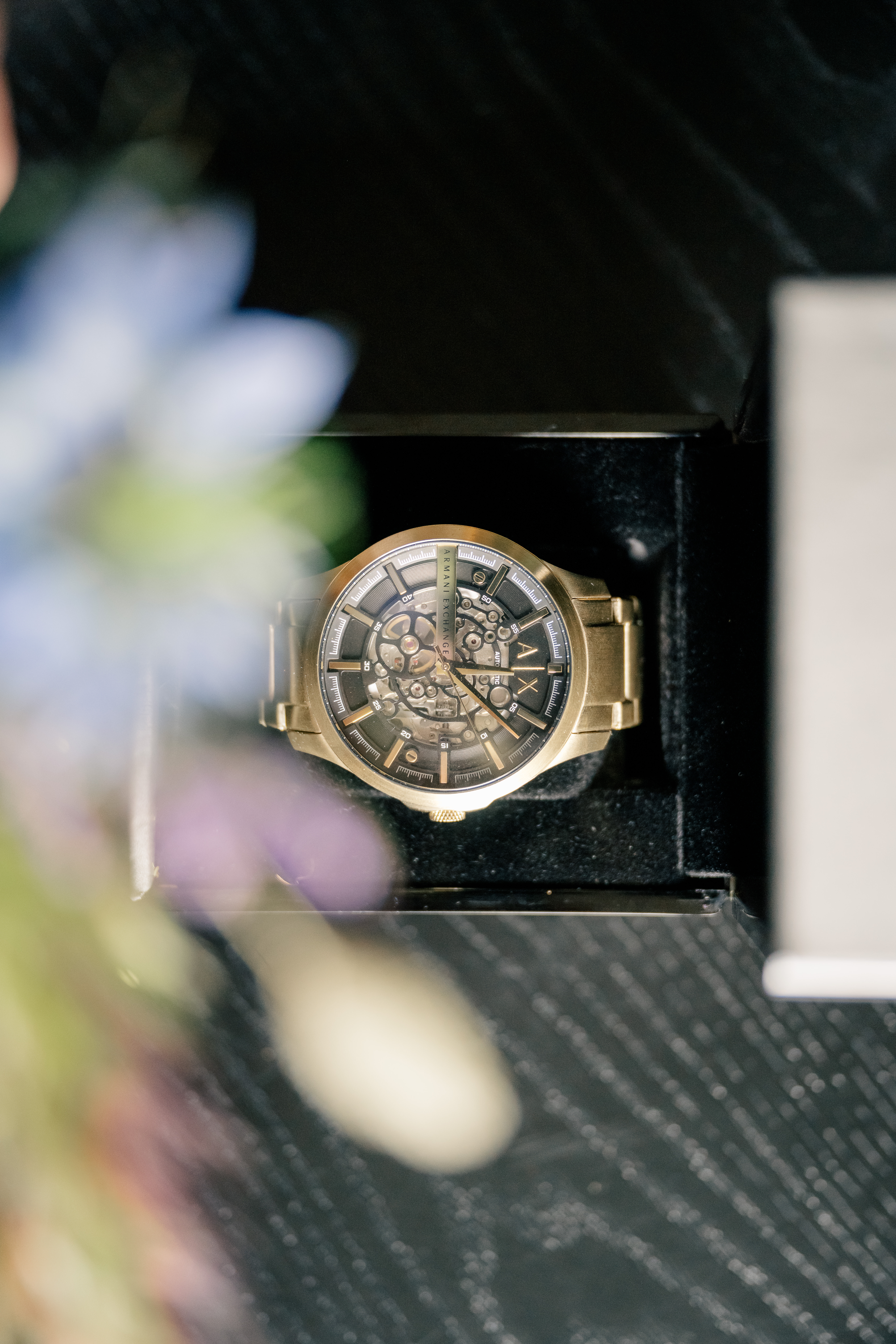 detail view of gold watch with visible gears in a box that is cream outside and black velvet inside with a view of purple flowers from the corsage by it