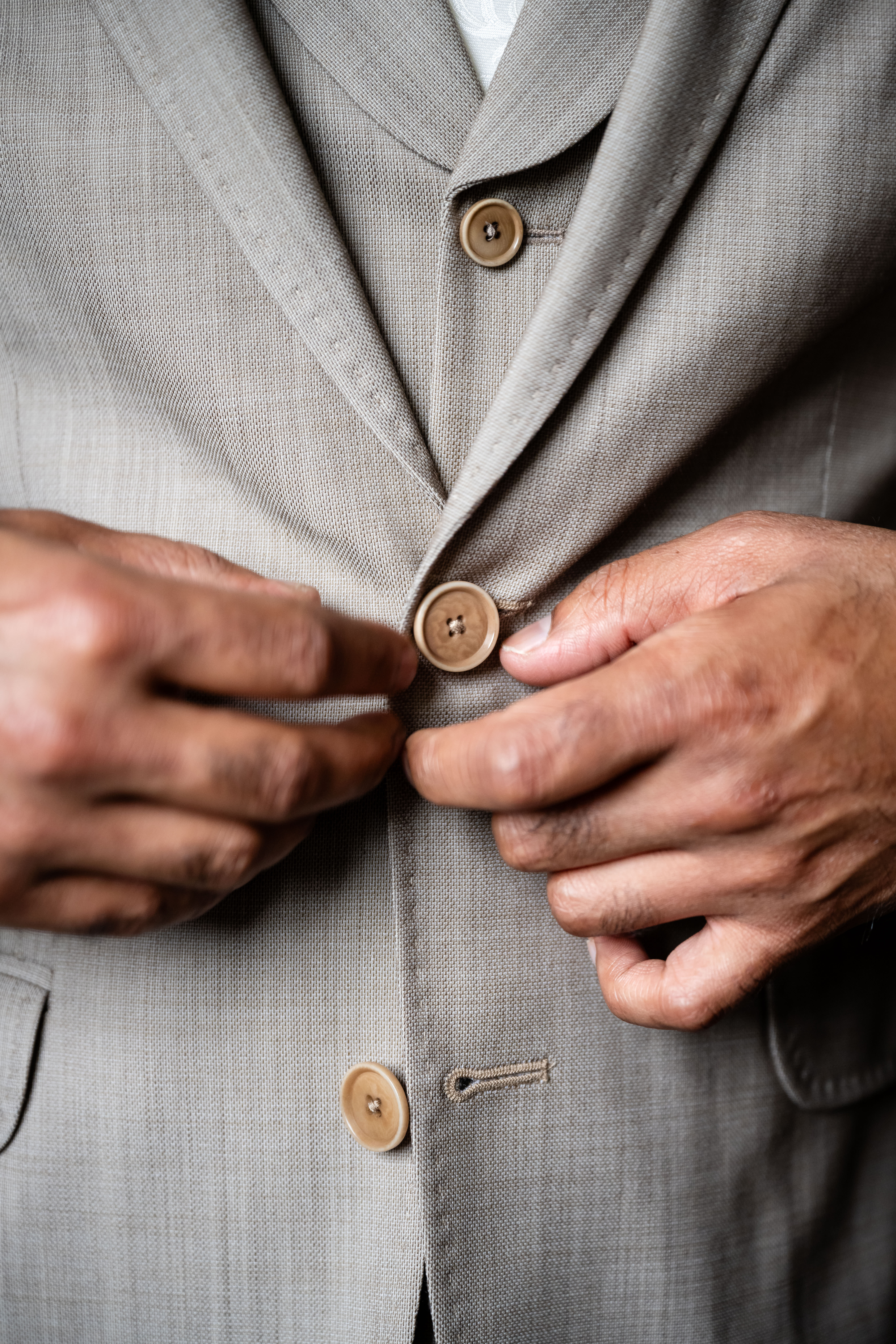 detail photo of a groom buttoning the wooden buttons of his beige wedding suit jacket up over his vest
