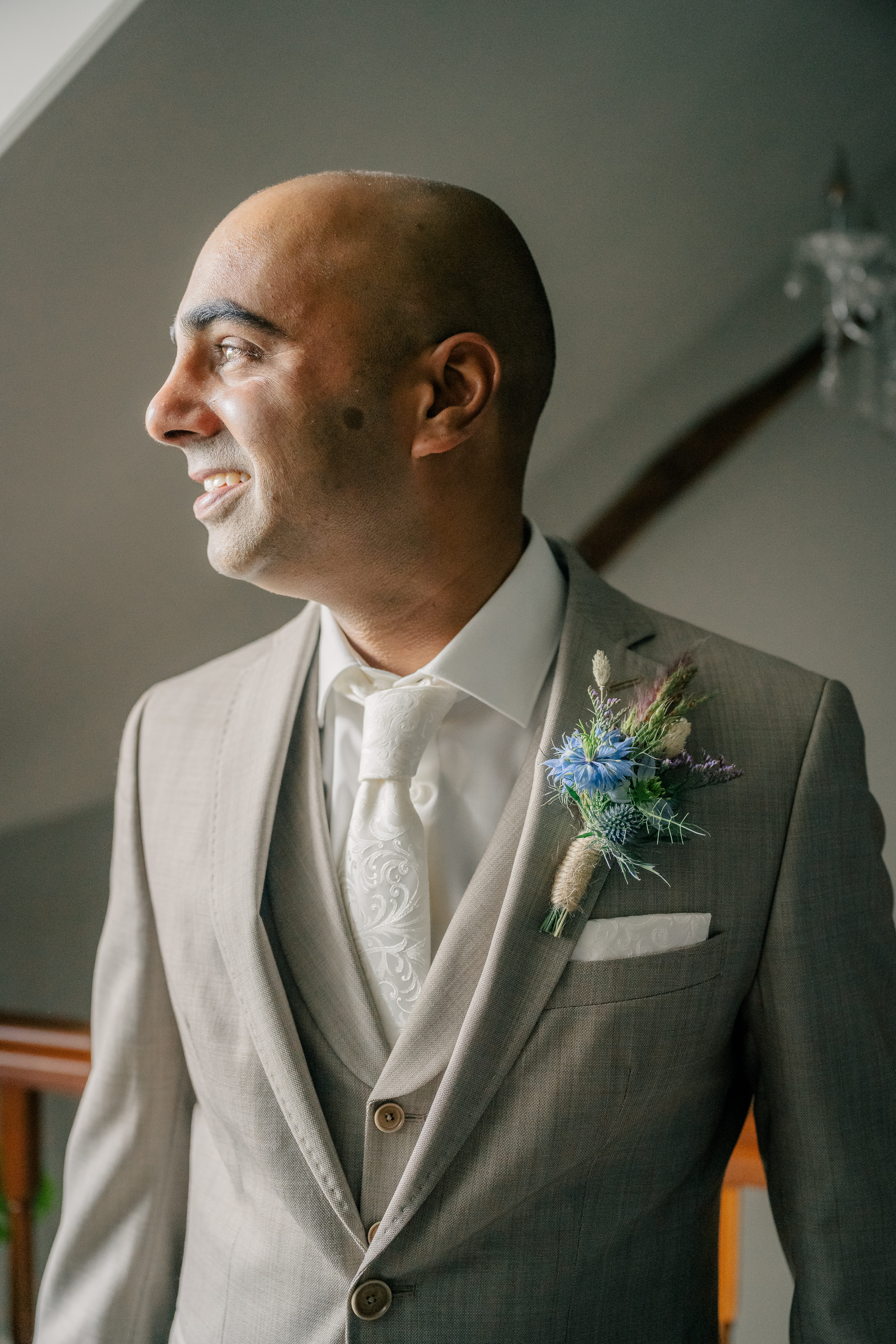 Profile photo of groom looking to the window after dressing in his beige three piece suit with white tie and corsage on his wedding day