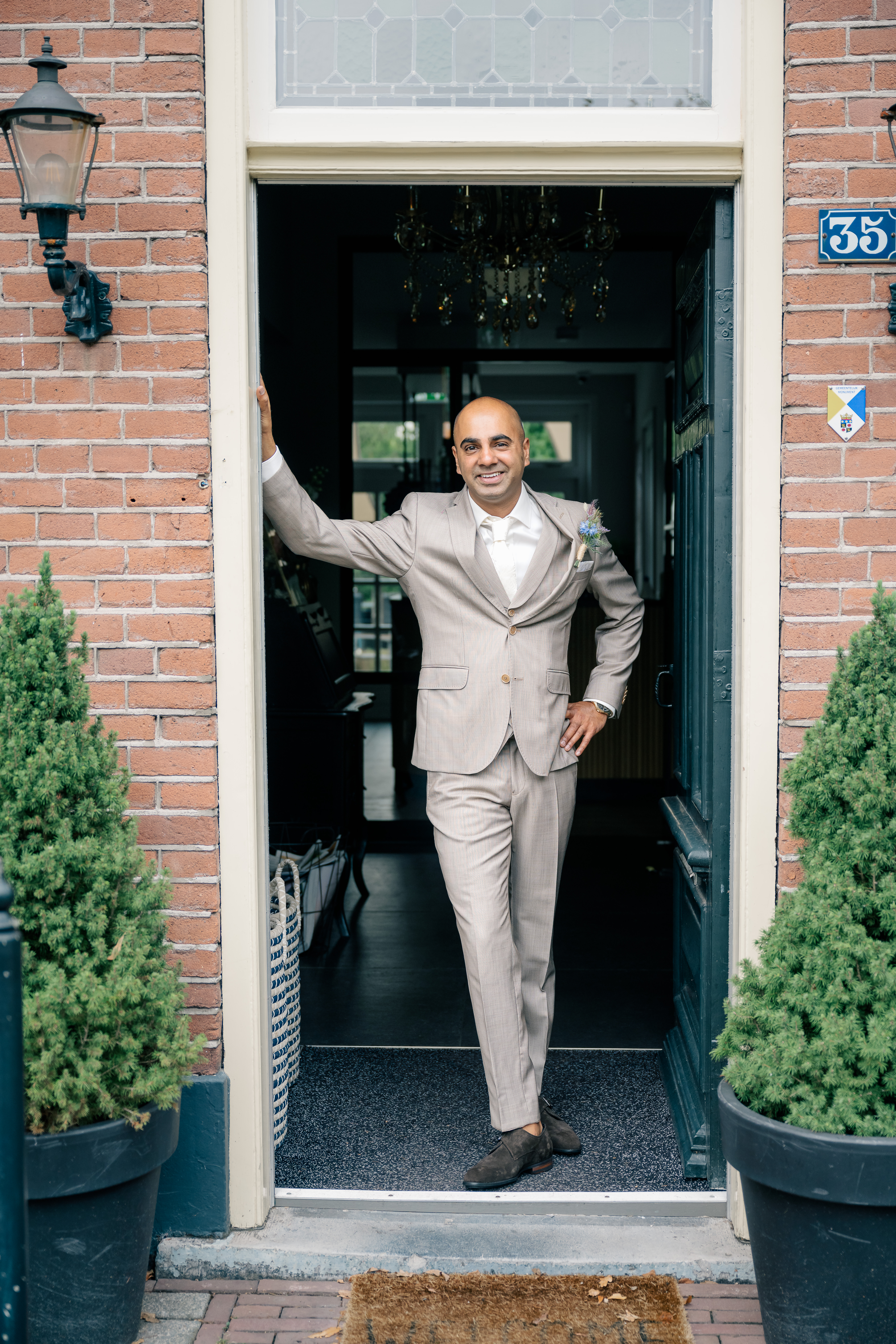 a groom stands wearing a beige suit and corsage leaning in the doorway of a brick monumental house in the Netherlands