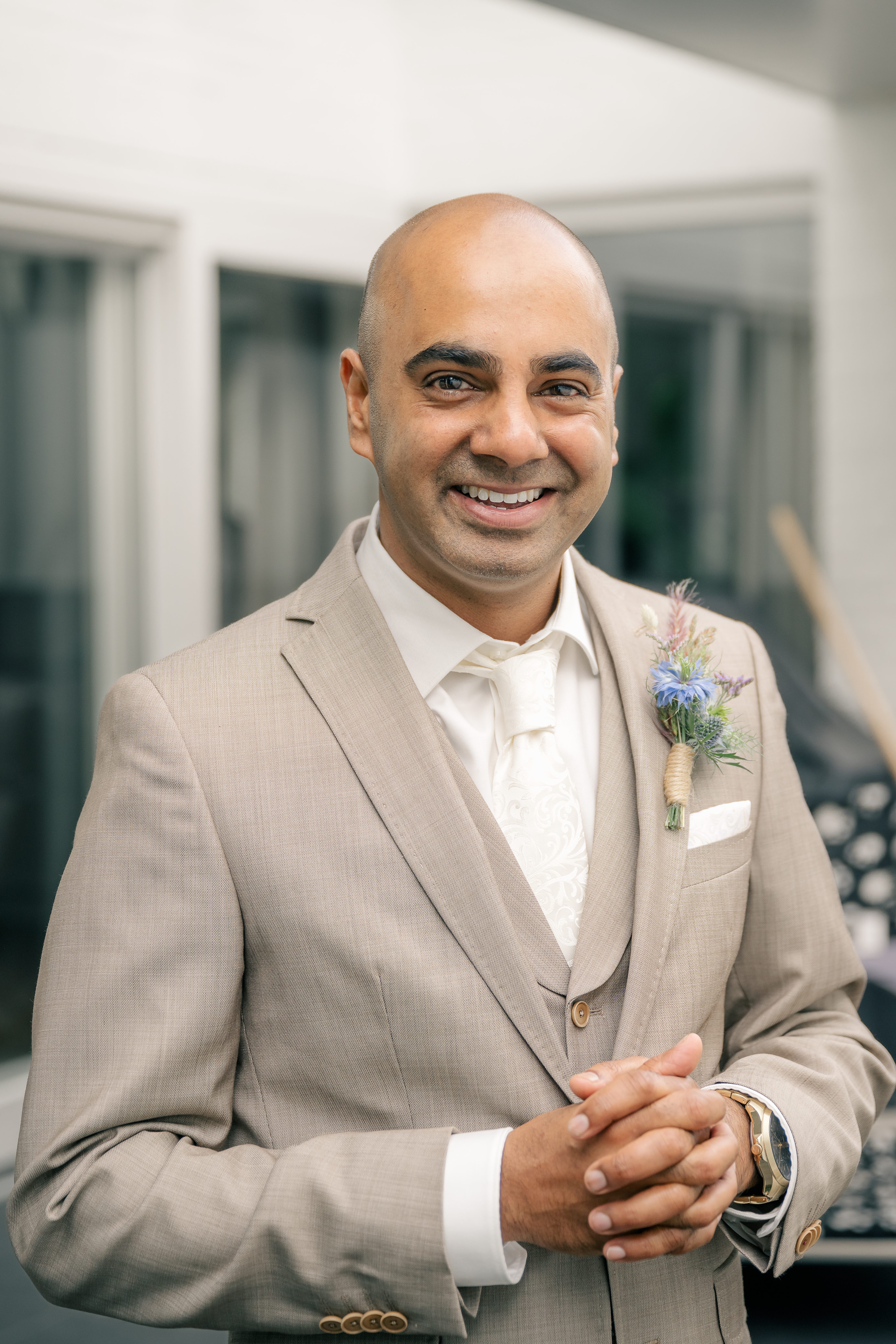 A groom in a beige suit looks excitedly at the camera as the bride walks up from behind for the First Look