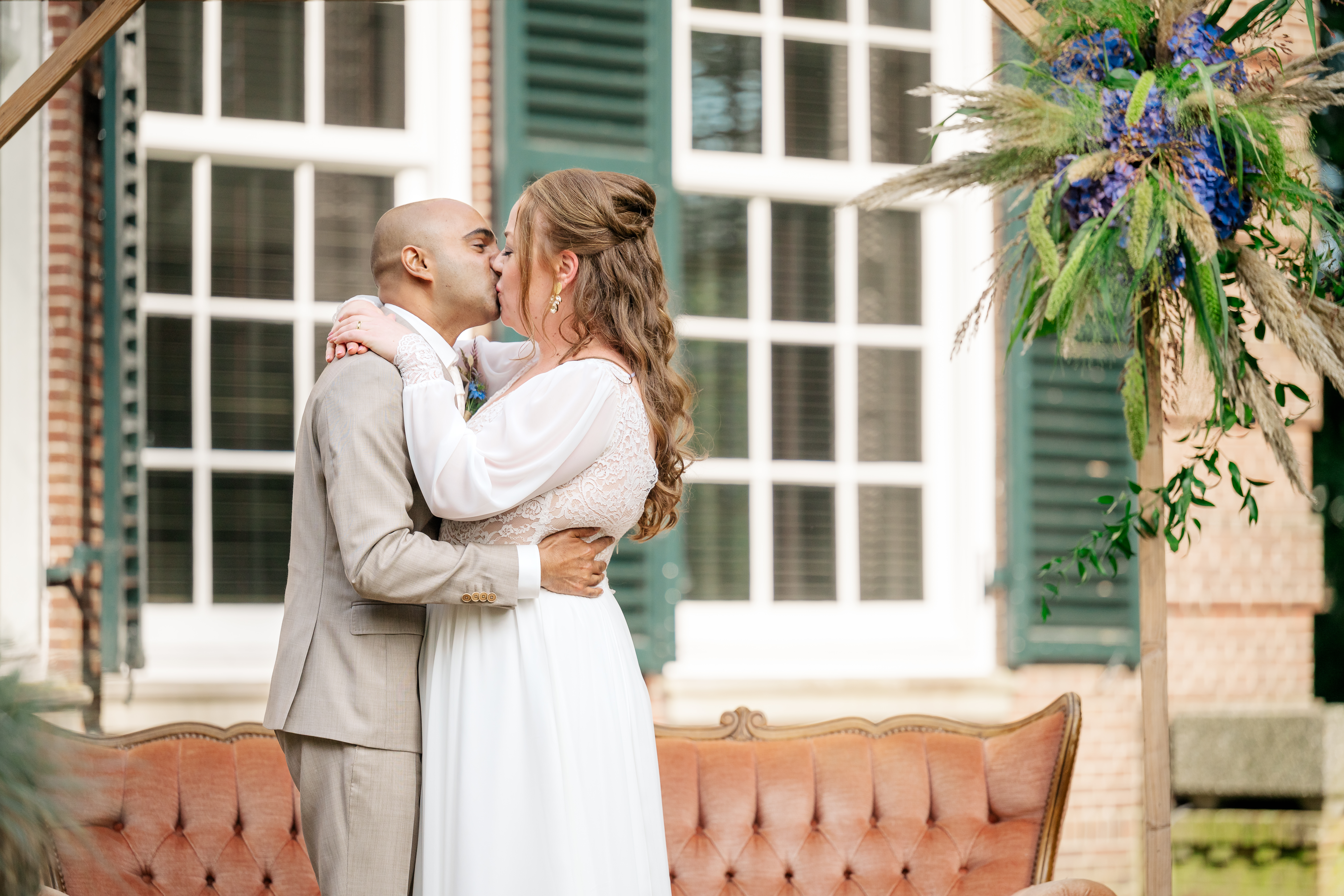 A bride in boho white wedding gown kisses a groom in beige suit under a wood and floral custom wedding arch in front of monumental villa and vintage sofa in Helmond