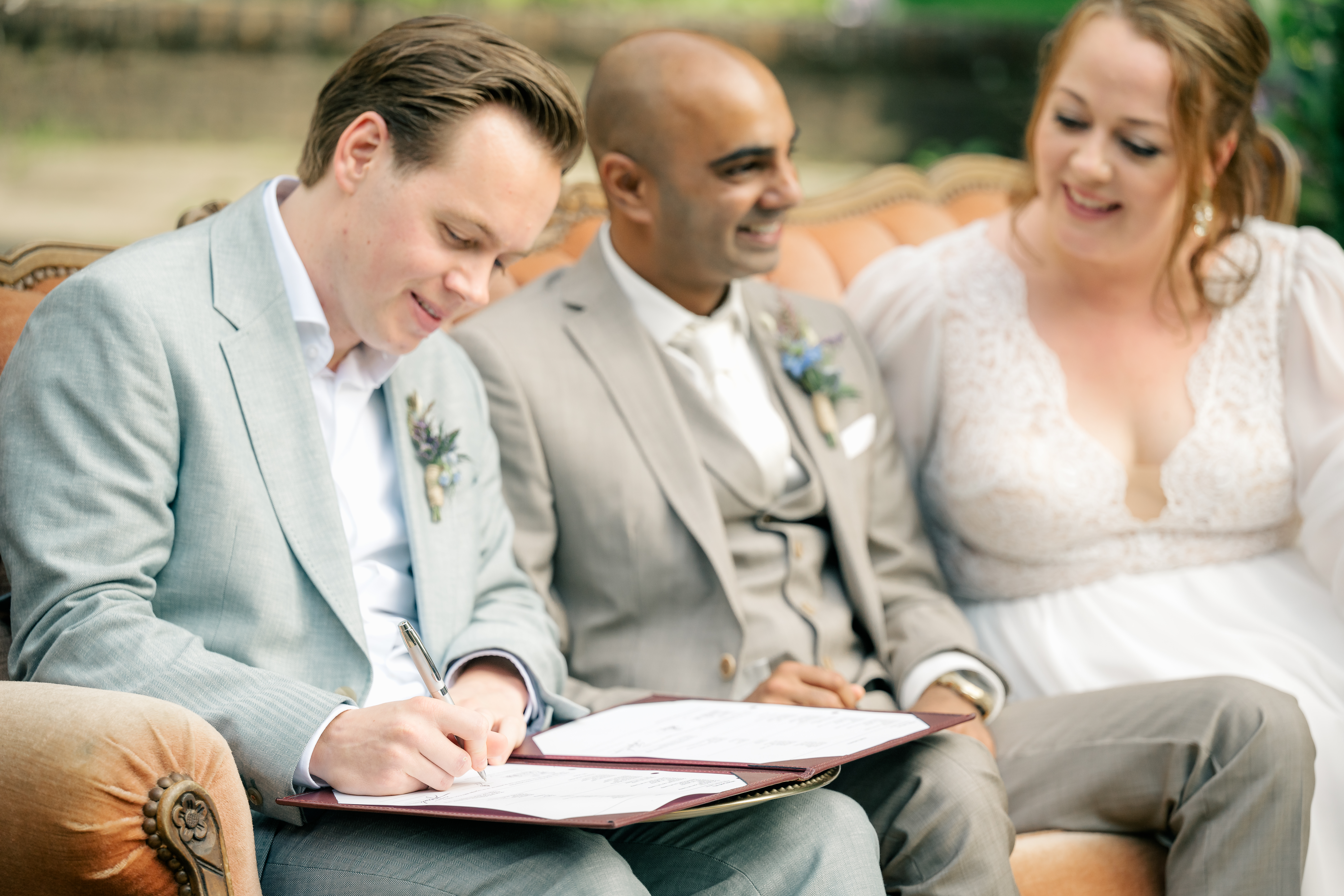 Three people are seated on a vintage ochre sofa outdoors. The man closest is a witness for the wedding and he signs the marriage record as the bride and groom look on