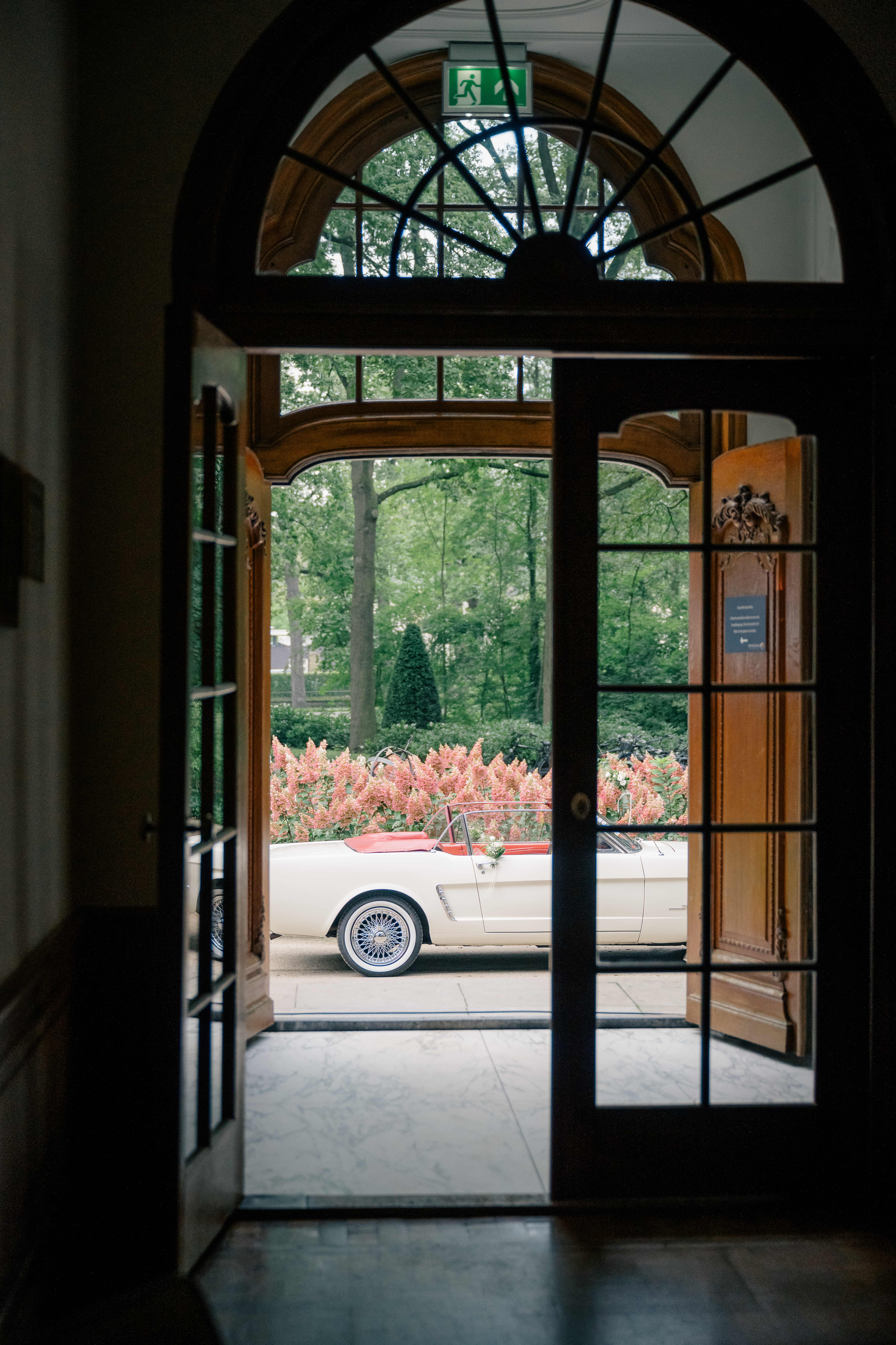 looking through wooden and glass doors of the villa in shadow to outside where the cream convertible wedding auto is parked on the circular driveway