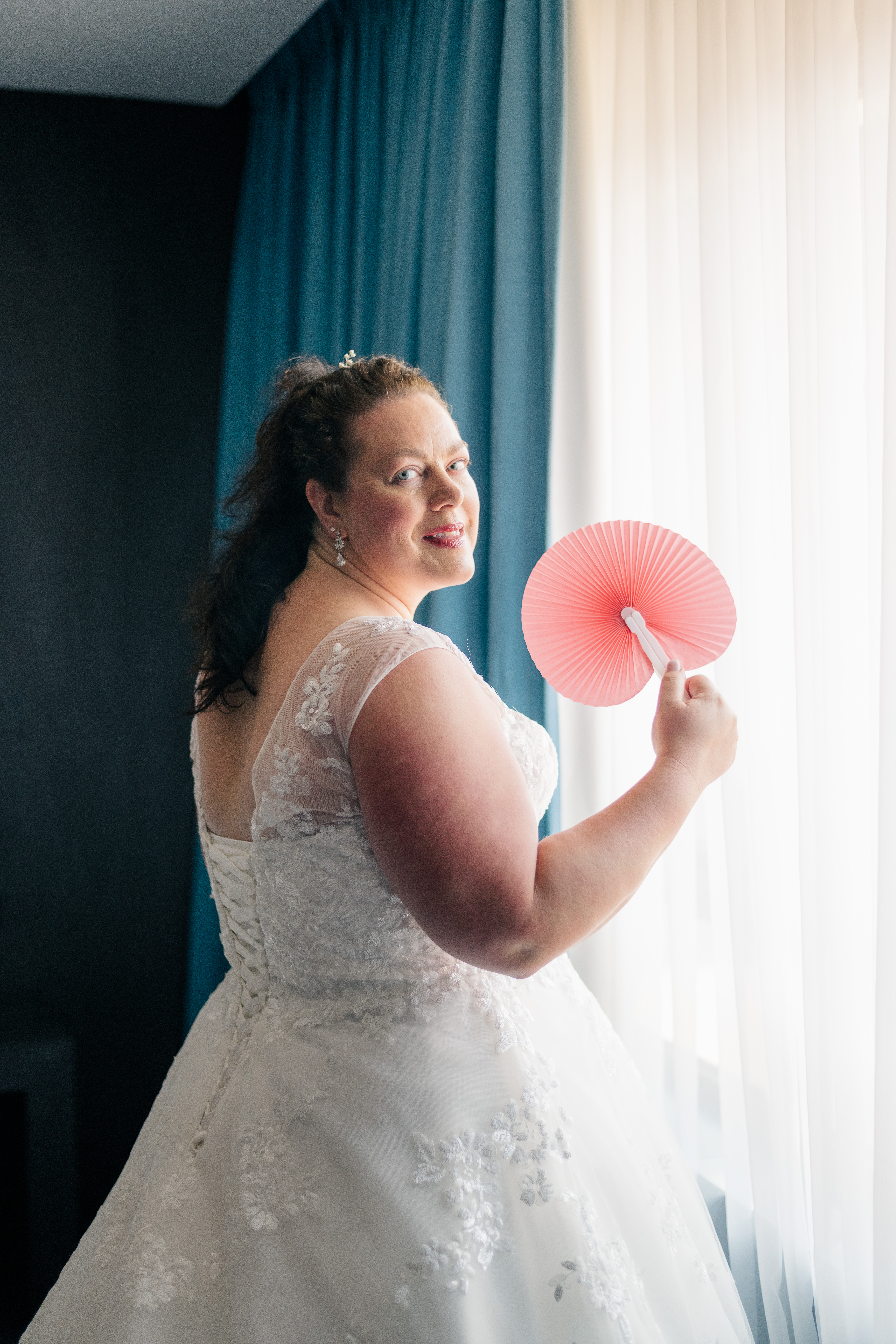 bride looking over her shoulder standing by a window with a coral hand fan in her hand