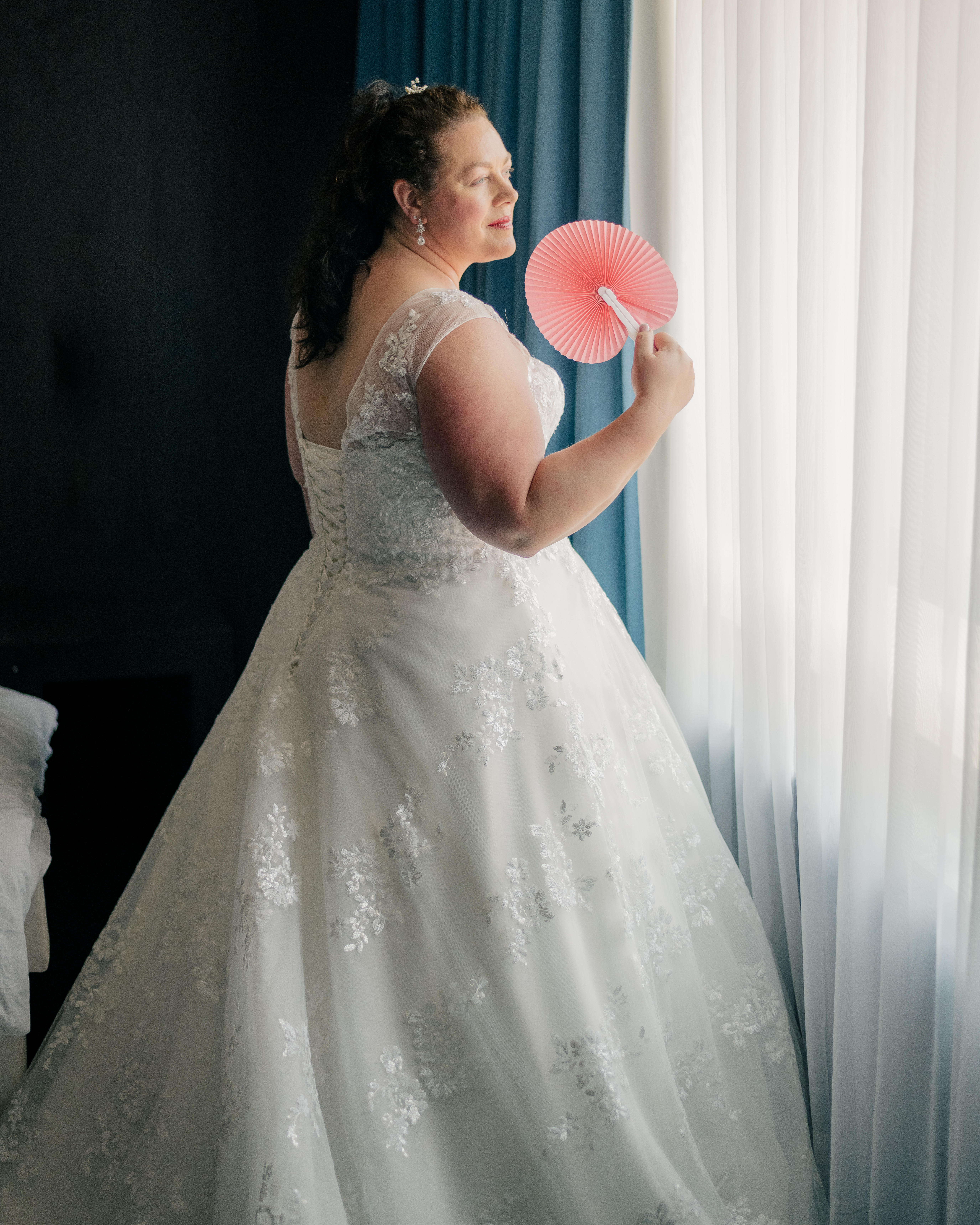 bridal portrait of a bride looking out a lightly curtained window holding a coral coloured hand fan