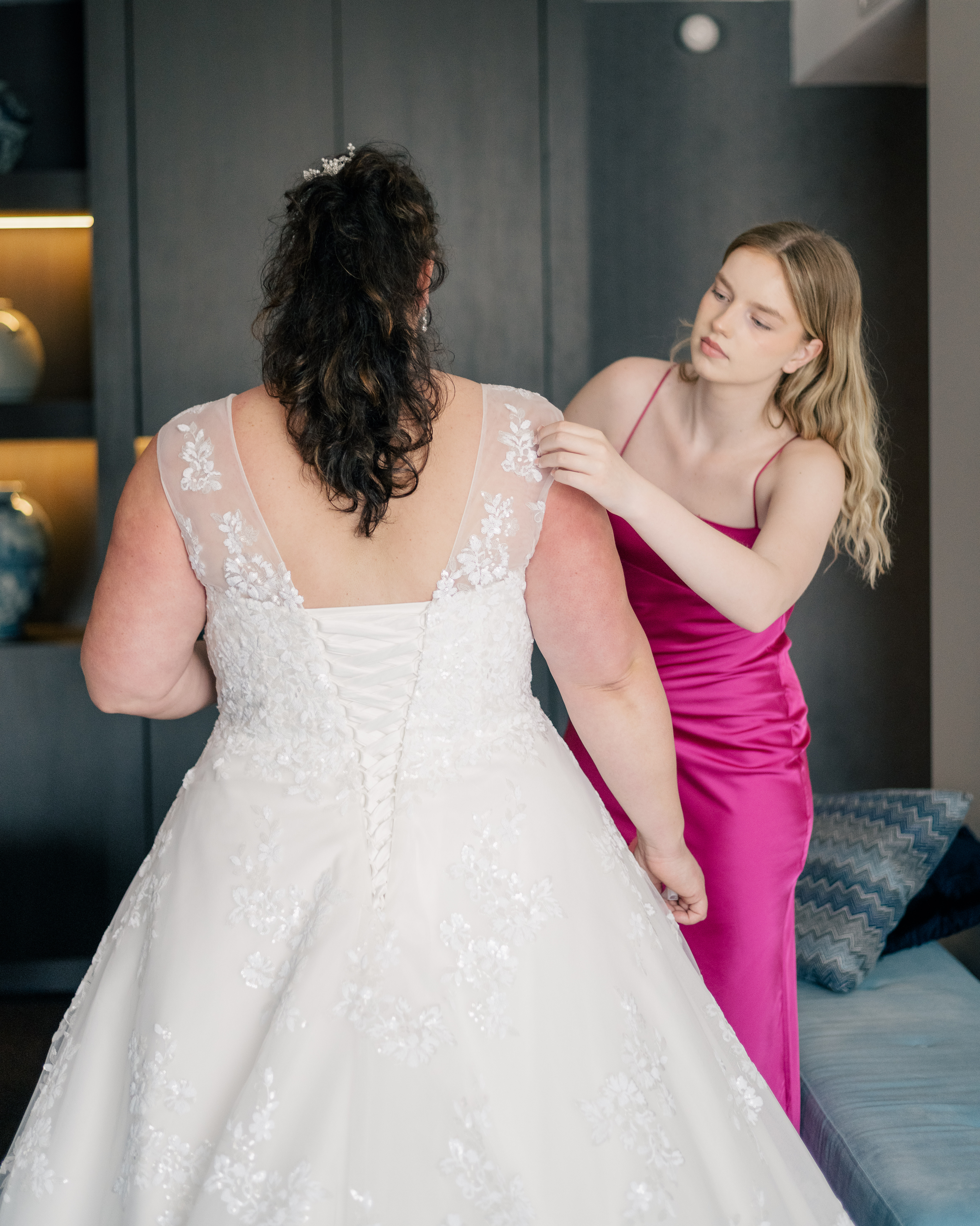 a bride getting ready in her white wedding dress with dark hair whilst her daughter with blond hair and a pink dress helps to her right