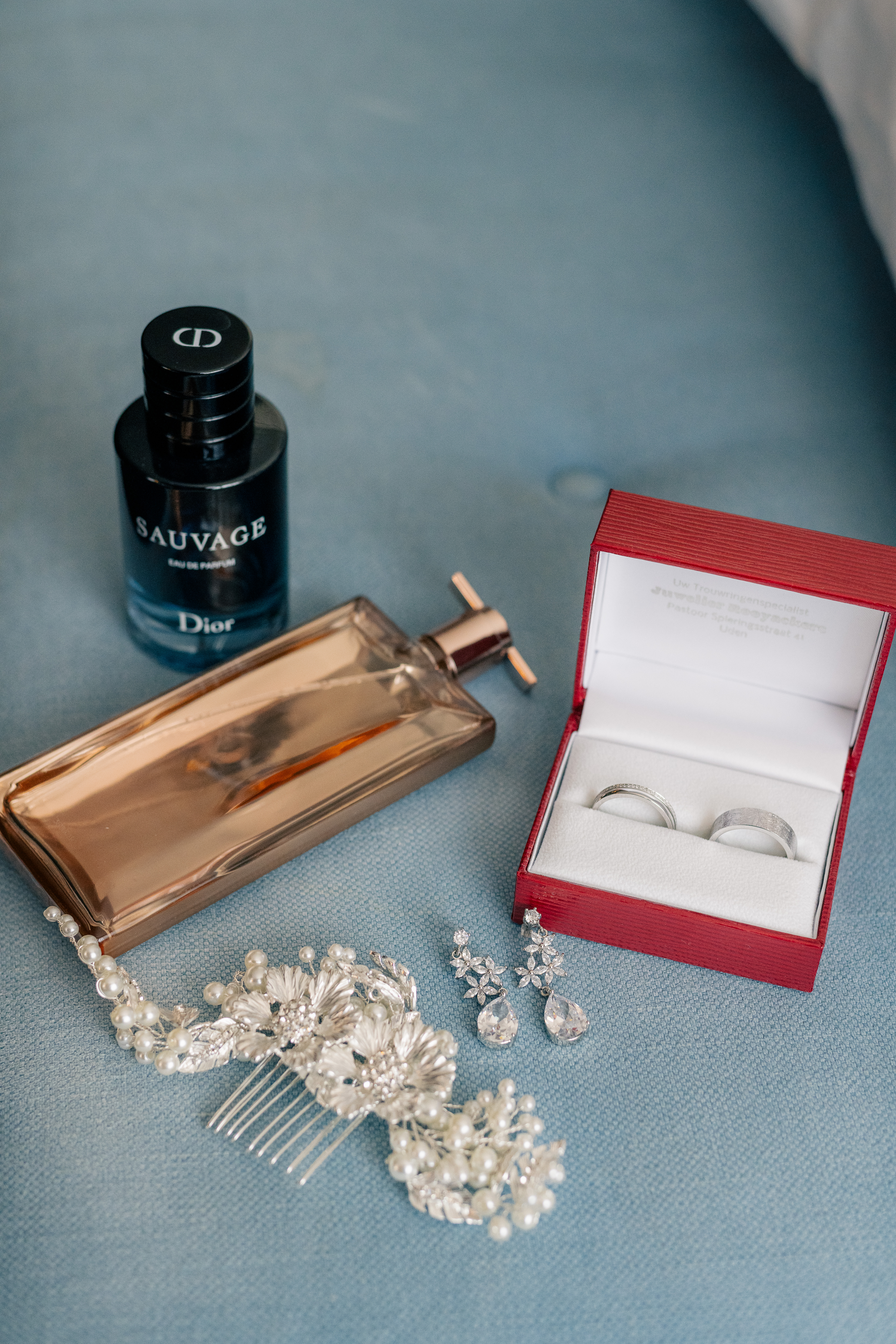 wedding details on a blue upholstered bench of a bridal hair comb, earrings, rings, perfume and cologne