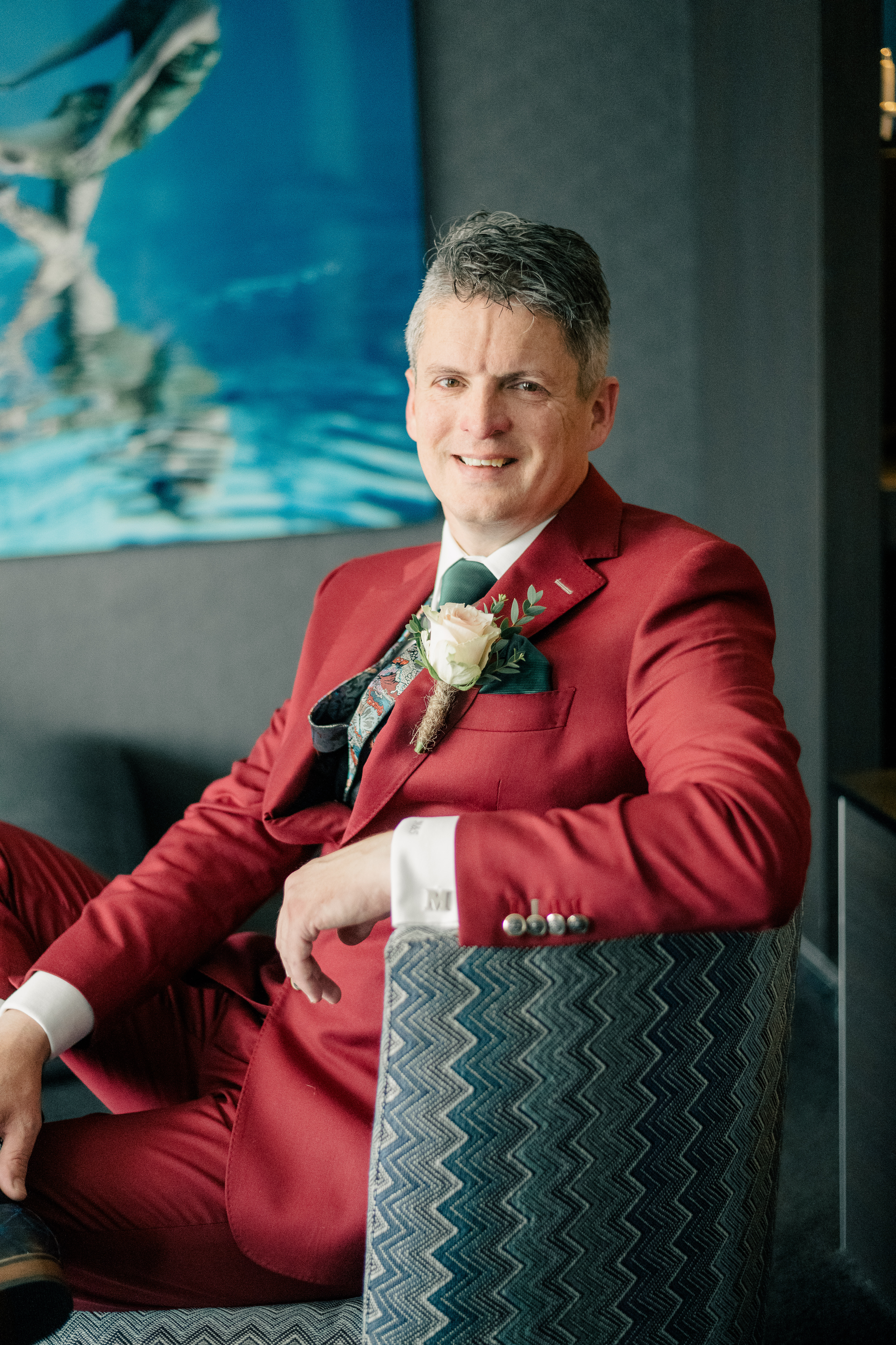 view of groom sitting in chair with leg crossed in a maroon coloured suit and cream coloured rose
