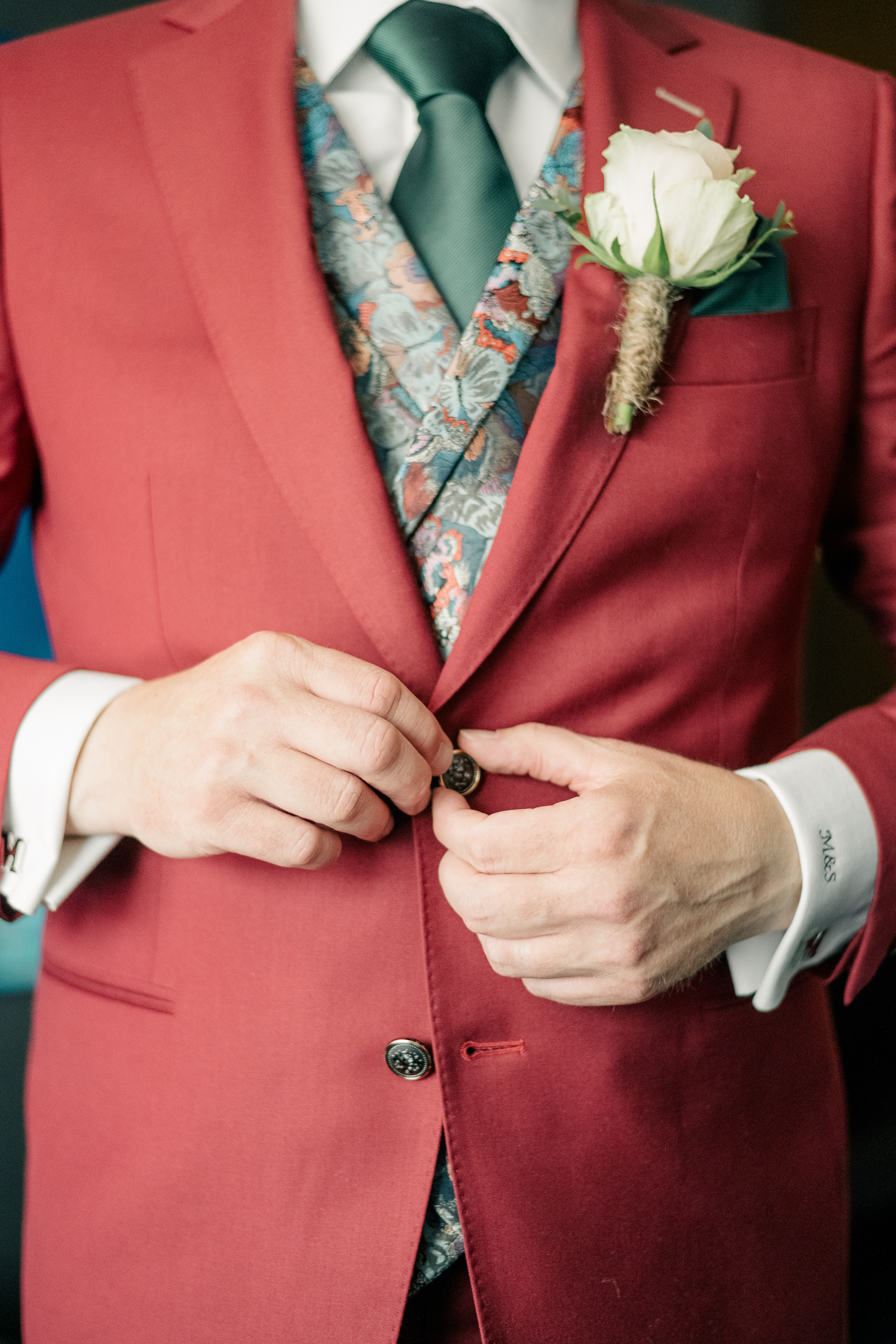 detail of groom buttoning maroon three piece suit with green tie, multi coloured vest, and cream coloured rose corsage