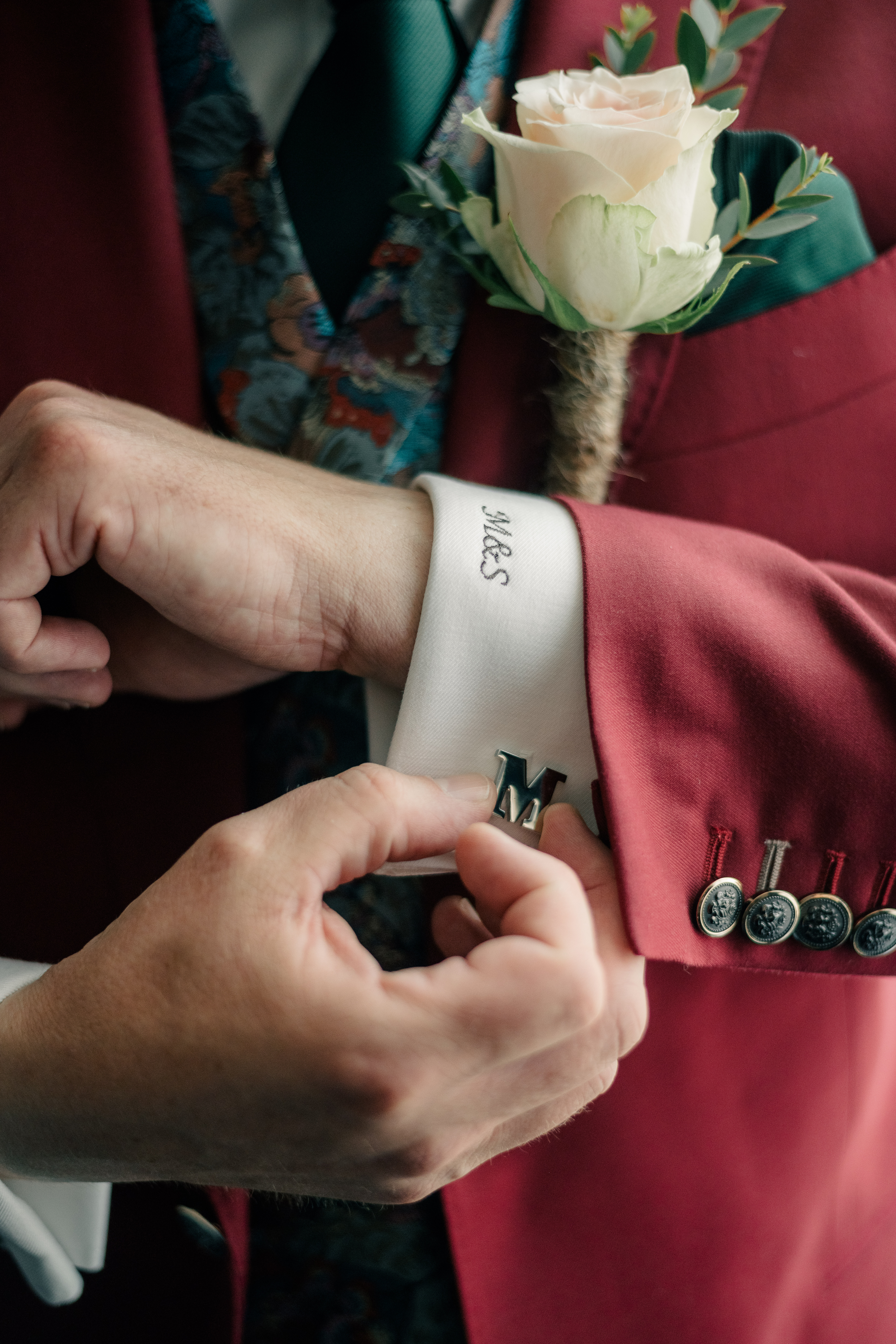 Detail of groom doing up his custom letter M cufflink with embroidered shirt cuff, three piece maroon suit, and pinkish cream rose