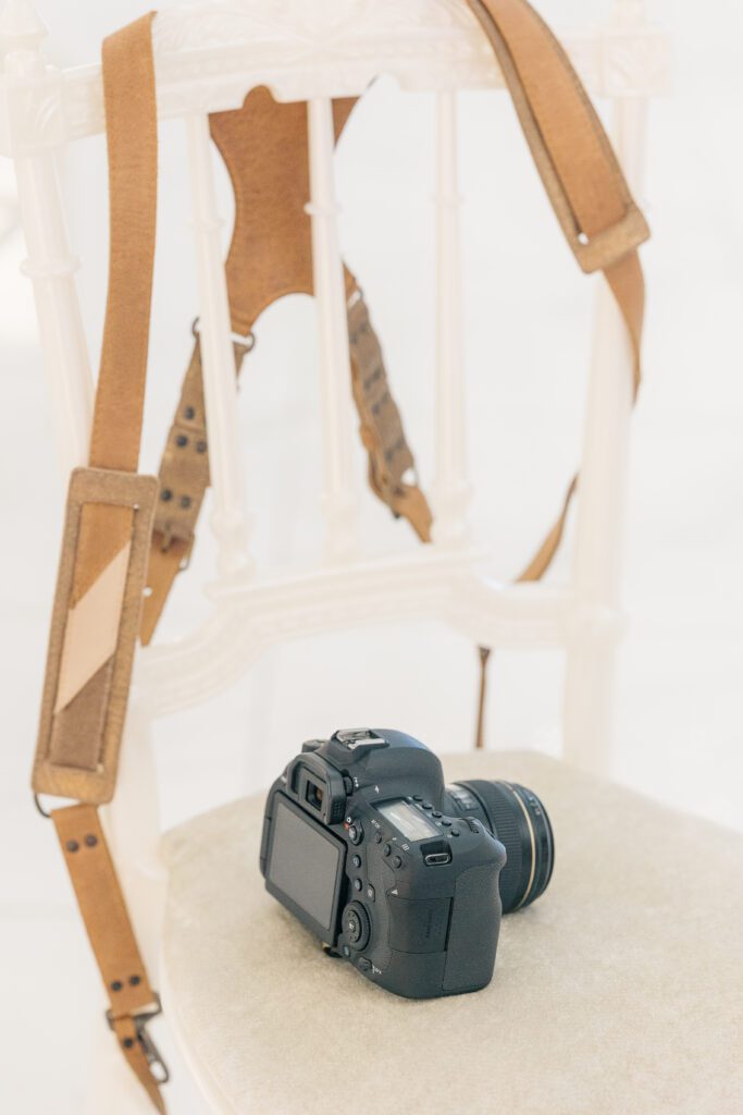 a camera and camera harness rest on a white wood and velour dining chair during a pause in photographing