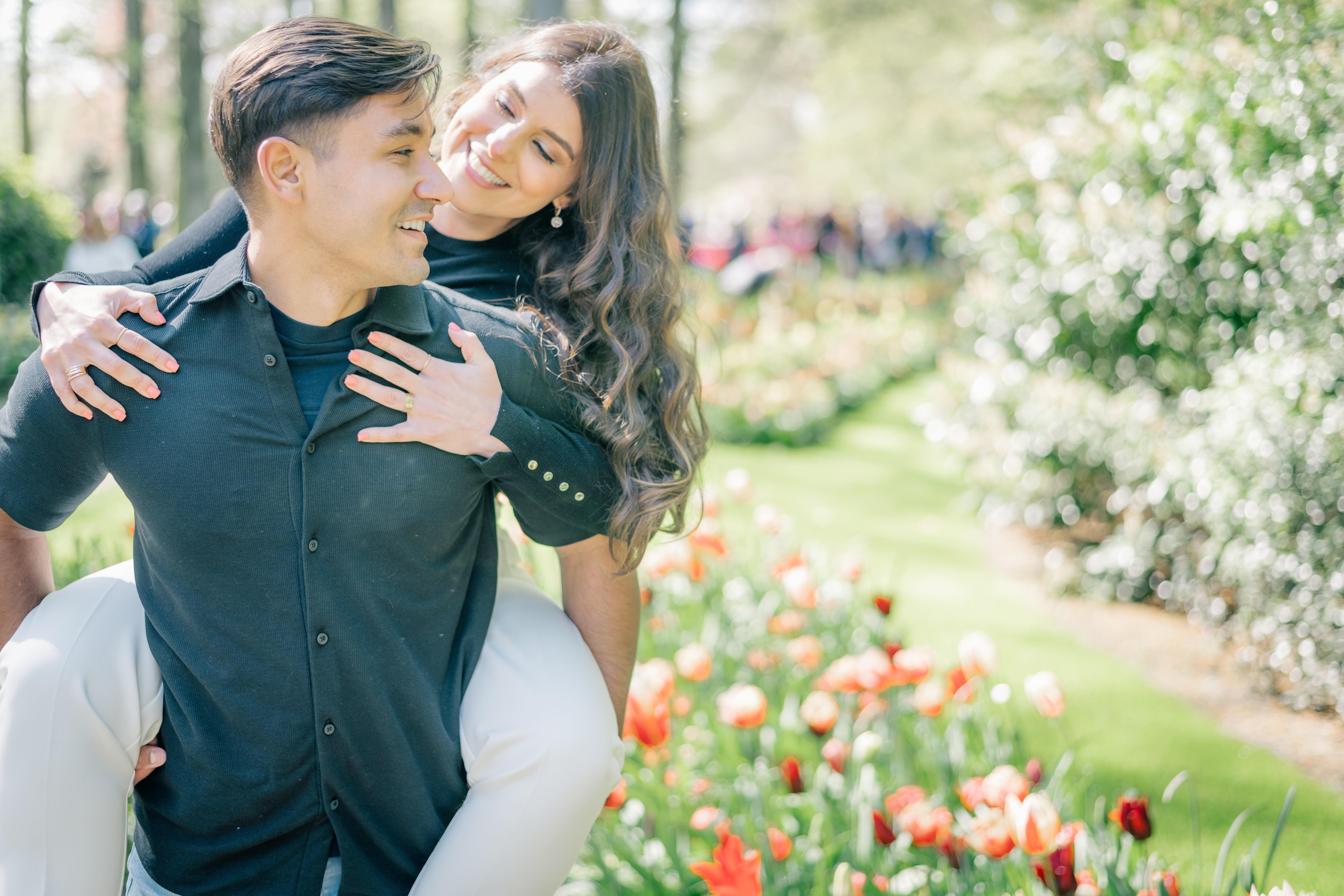 A woman rides on the back of a man and looks at him smiling while he looks back at her smiling.  They are standing in a garden full of tulips and other blooms