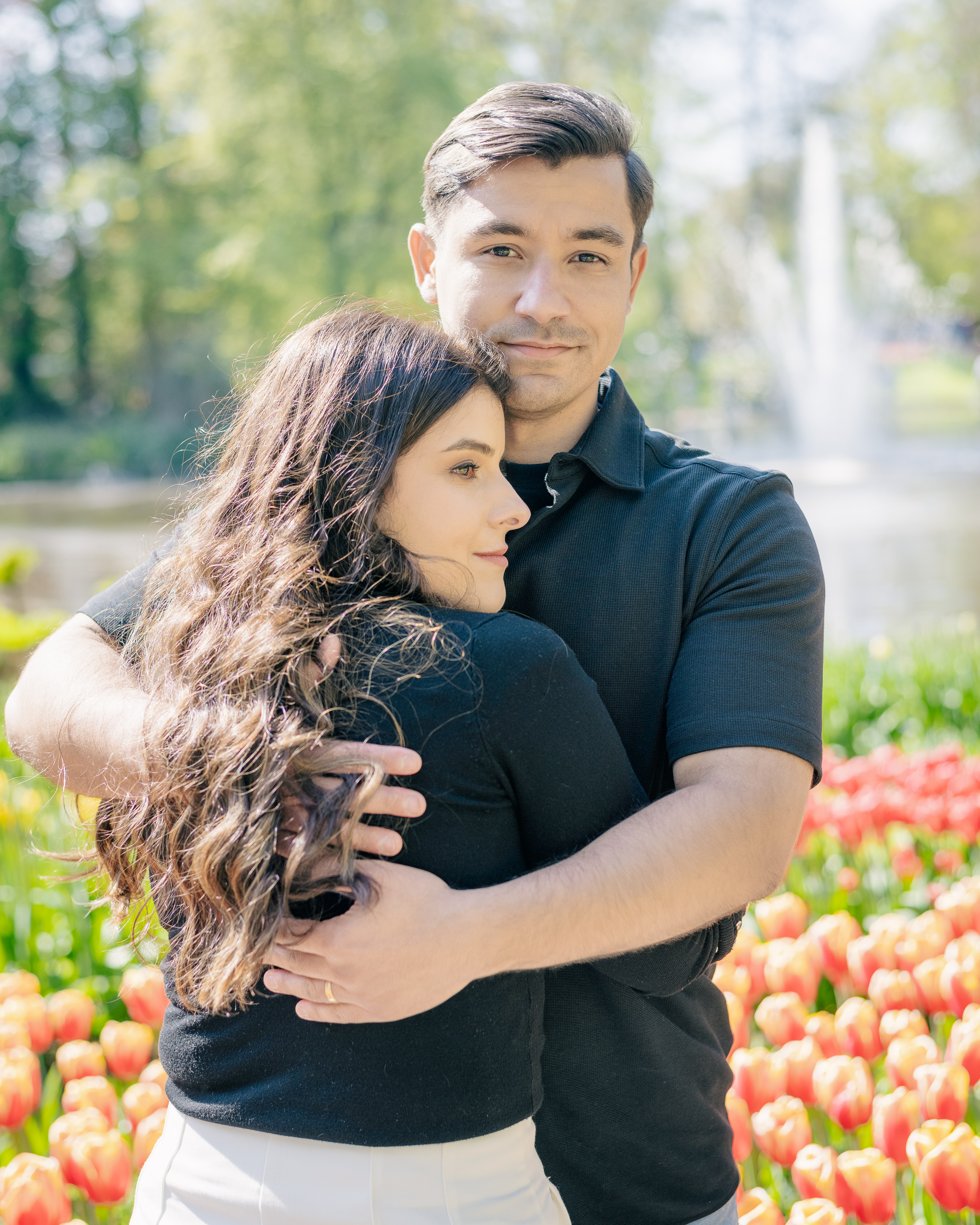 A man hugs his wife close while looking straight at the camera.  The wife looks softly to the left.  There are tulips and a fountain behind them