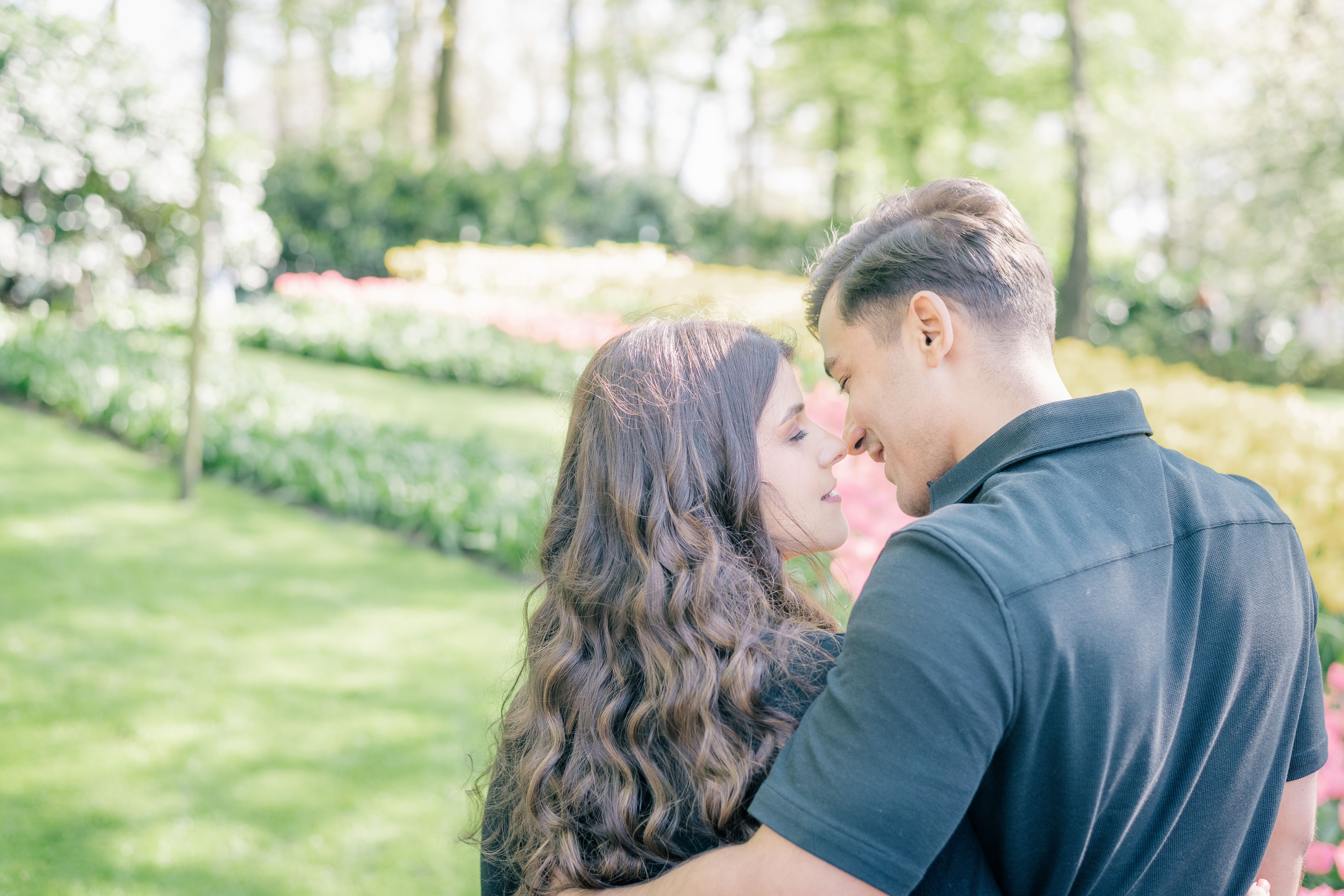 A man stands next to a woman with his arm around her waist.  They are standing with their eyes closed in front of blooming tulip beds and trees and are just about to kiss.  