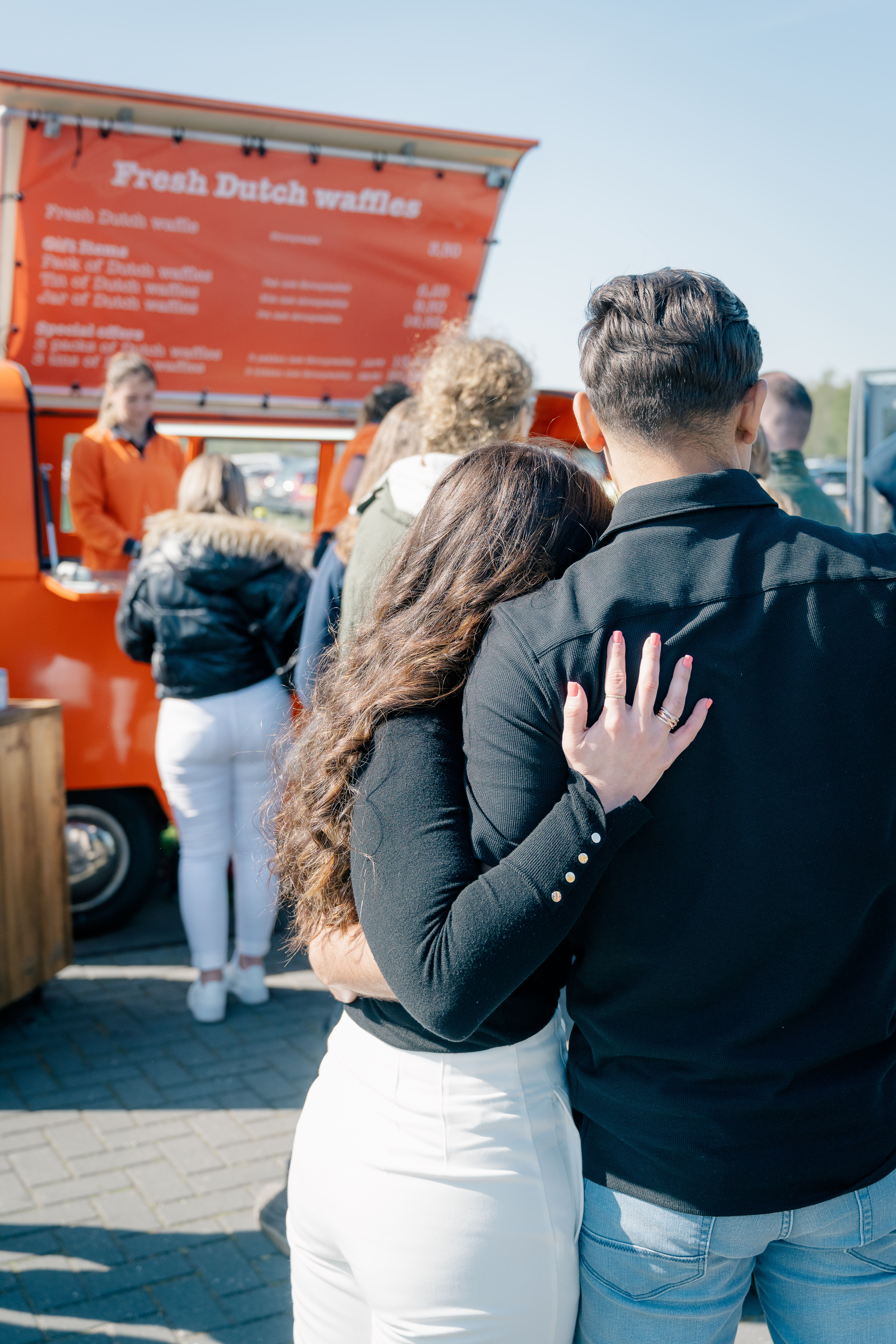 A man and woman stand with their arms around each other as they wait in line at an orange van selling fresh Dutch waffles