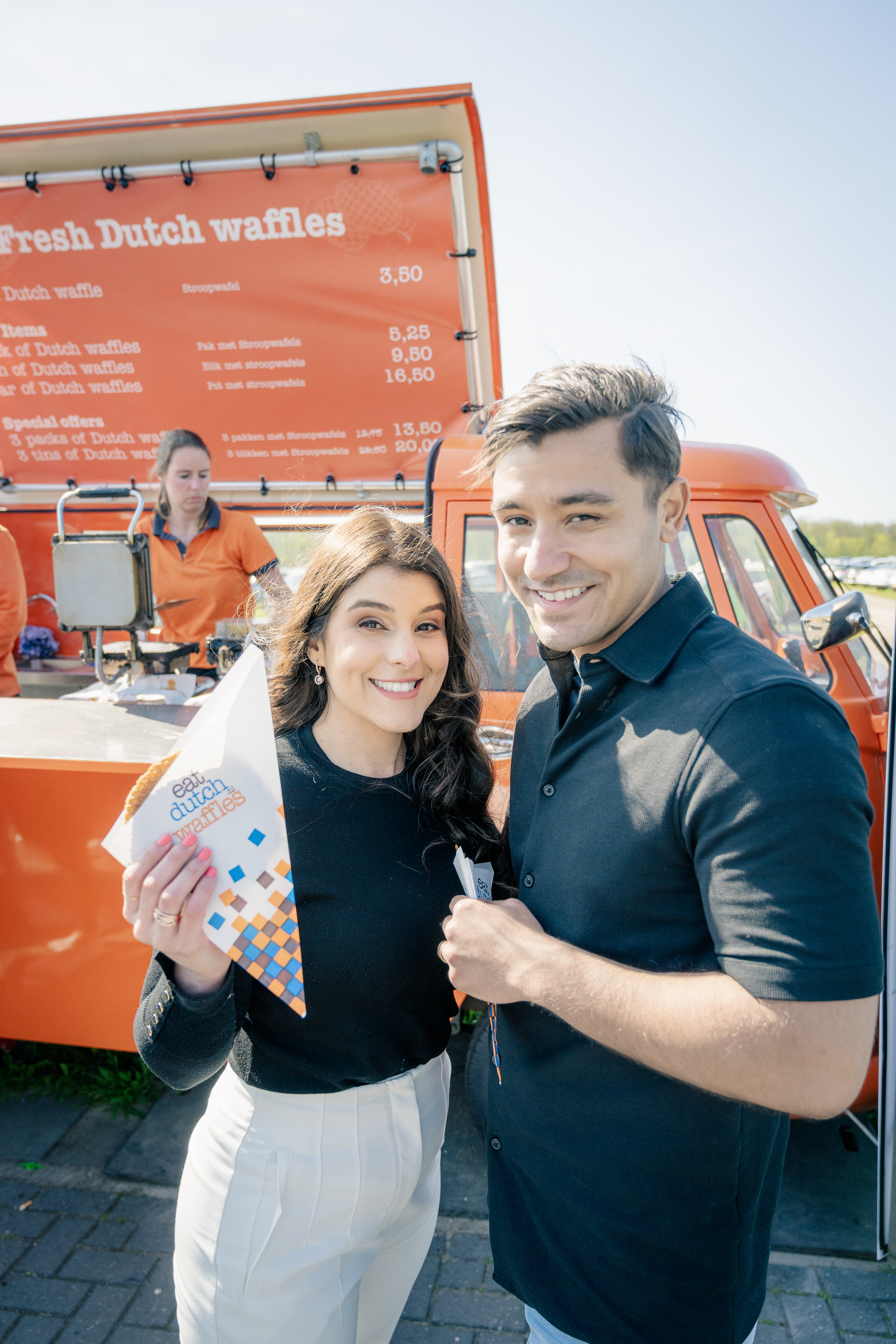 A woman and a man stand and smile at the camera in front of the orange Fresh Dutch waffles van after buying their stroopwafels 