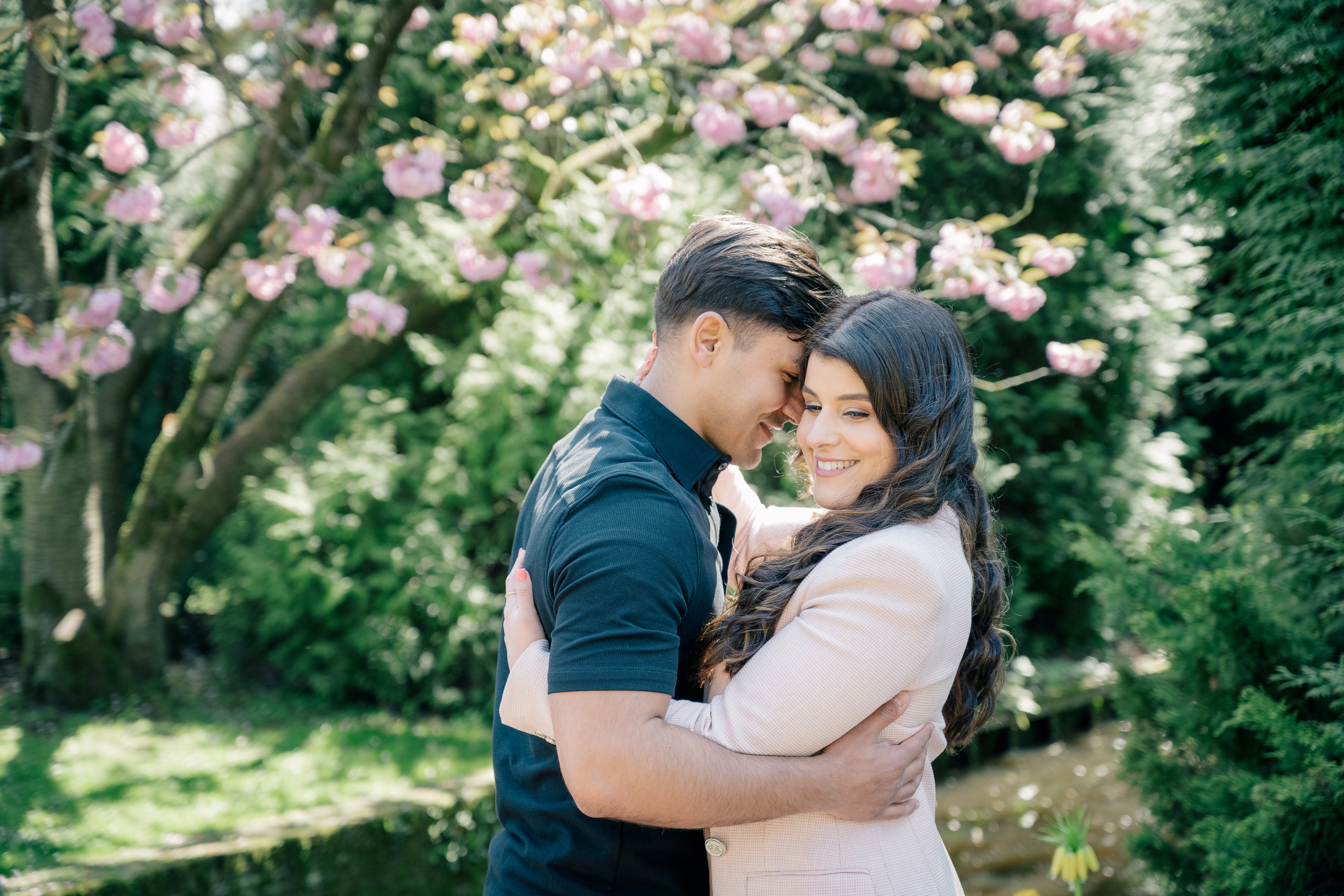A husband and wife share a loving embrace under a blooming magnolia tree in a sun dappled Keukenhof