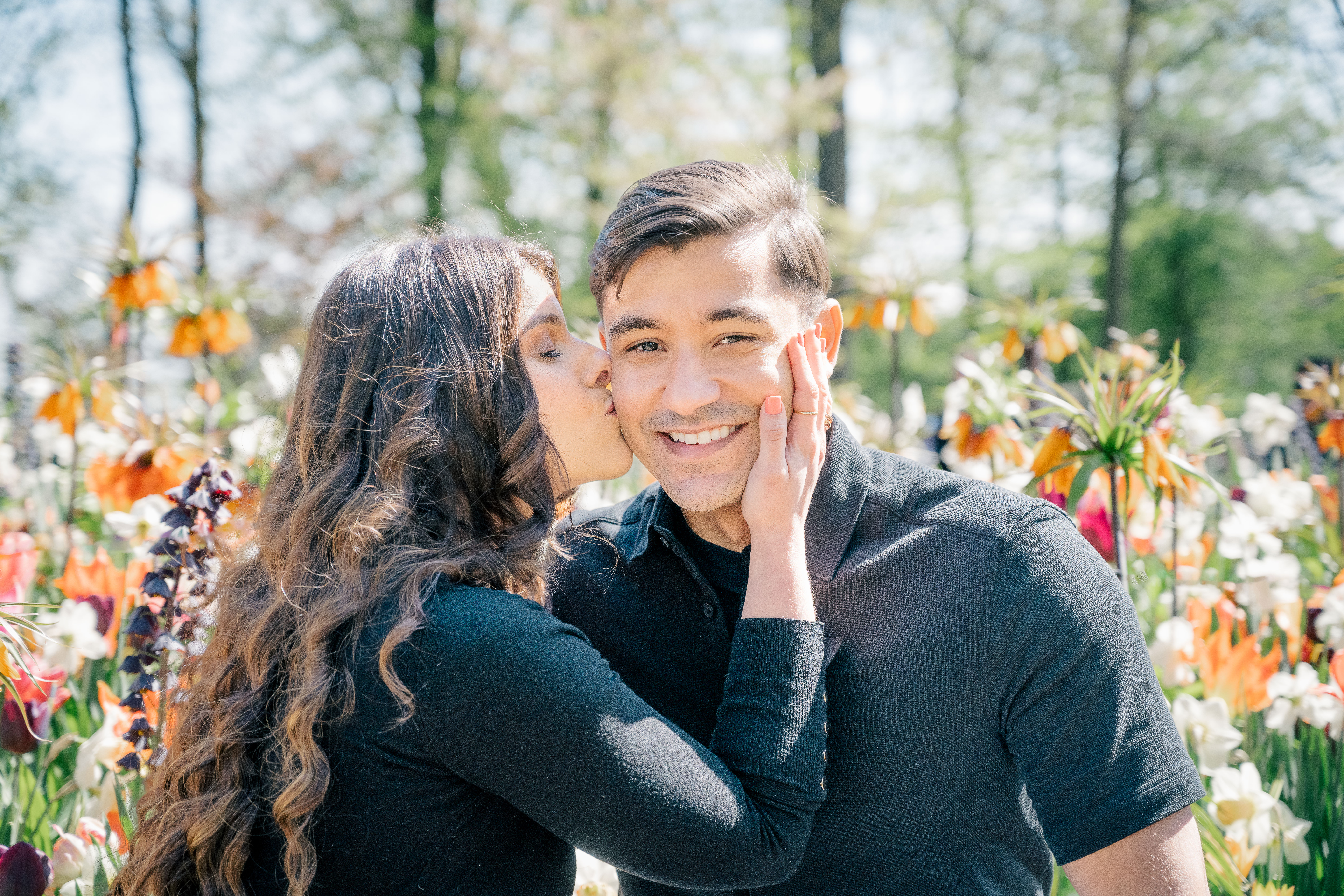 A couple both wearing black shirts and with dark brown hair sit in front of a colourful flowerbed in the Keukenhof.  The husband looks at the camera smiling and the wife kisses his cheek with her right hand on his opposite cheek and her eyes closed. 