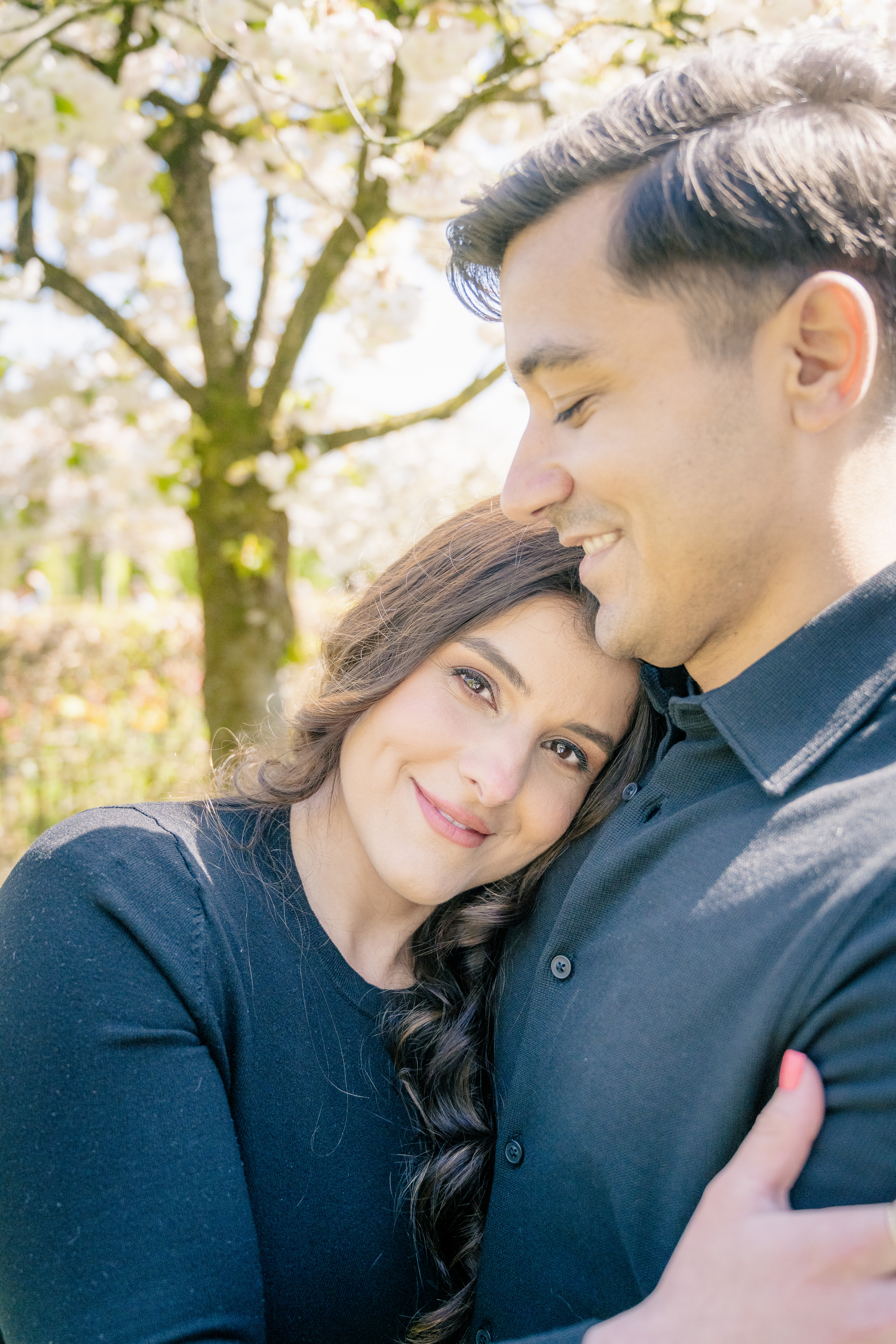 a woman lays her head on the chest of her man.  They are wearing black shirts and have dark brown hair.  The husband smiles down at her and she smiles at the camera