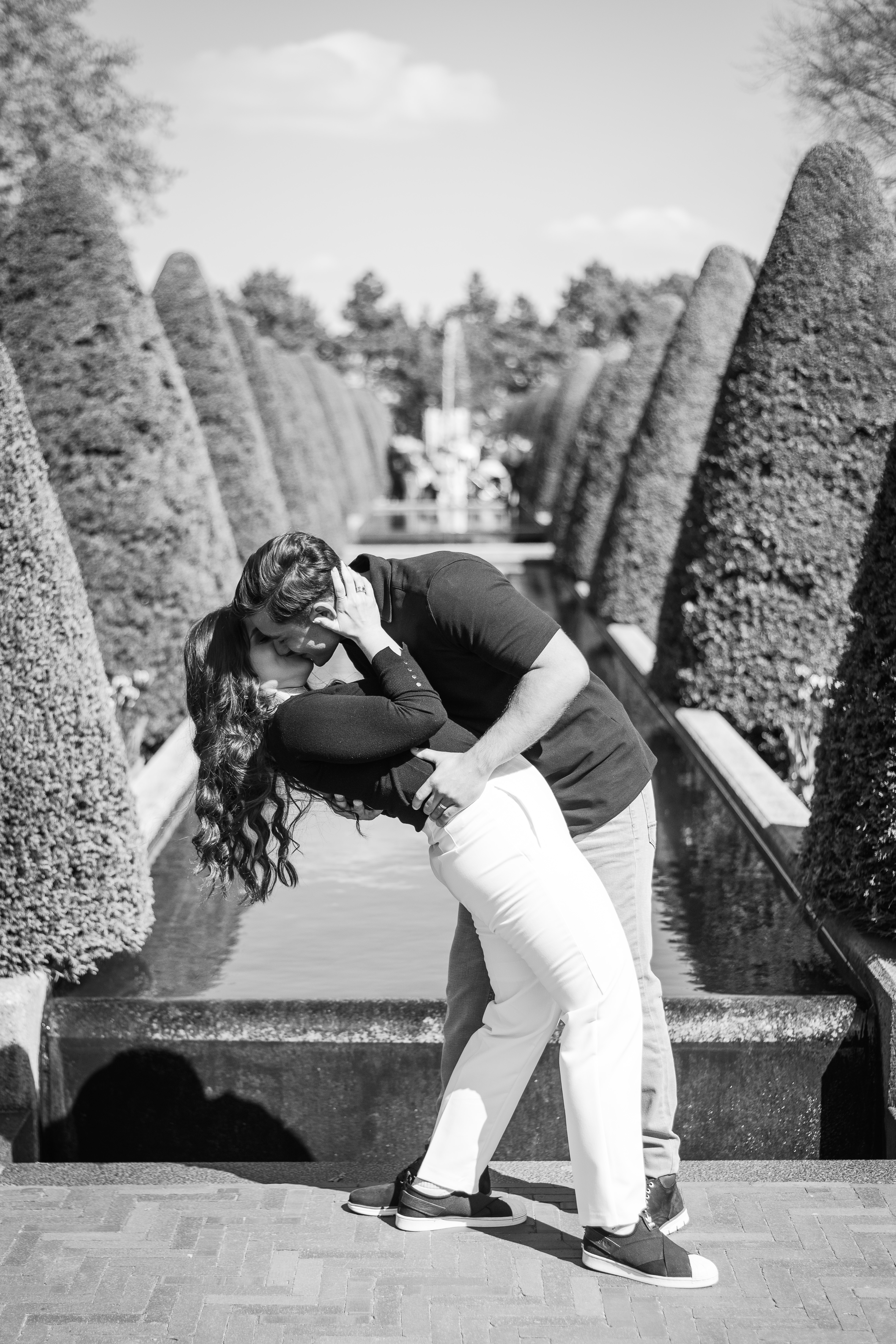 Black and white image of a man dipping a woman back to kiss her in front of a long row of trees and a water fountain