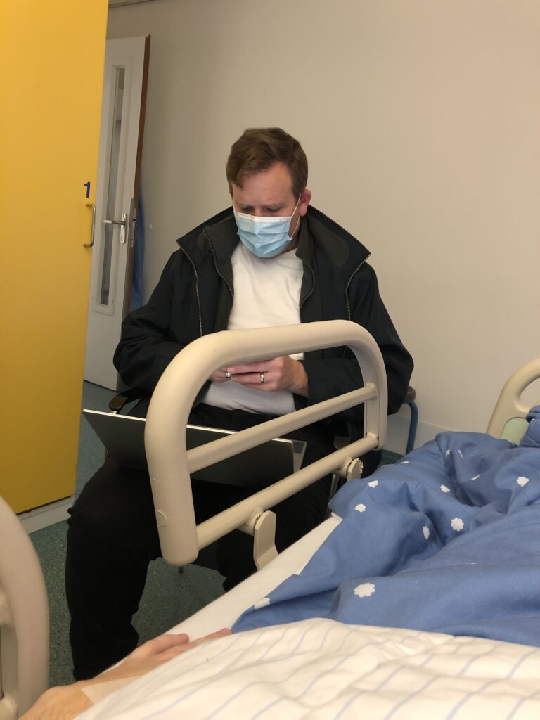 a man sits next to a hospital bed wearing a mask and working on a laptop