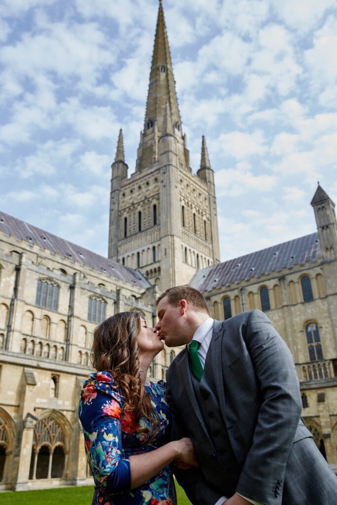A husband and wife kiss in Norwich Cathedral with the spire in the background