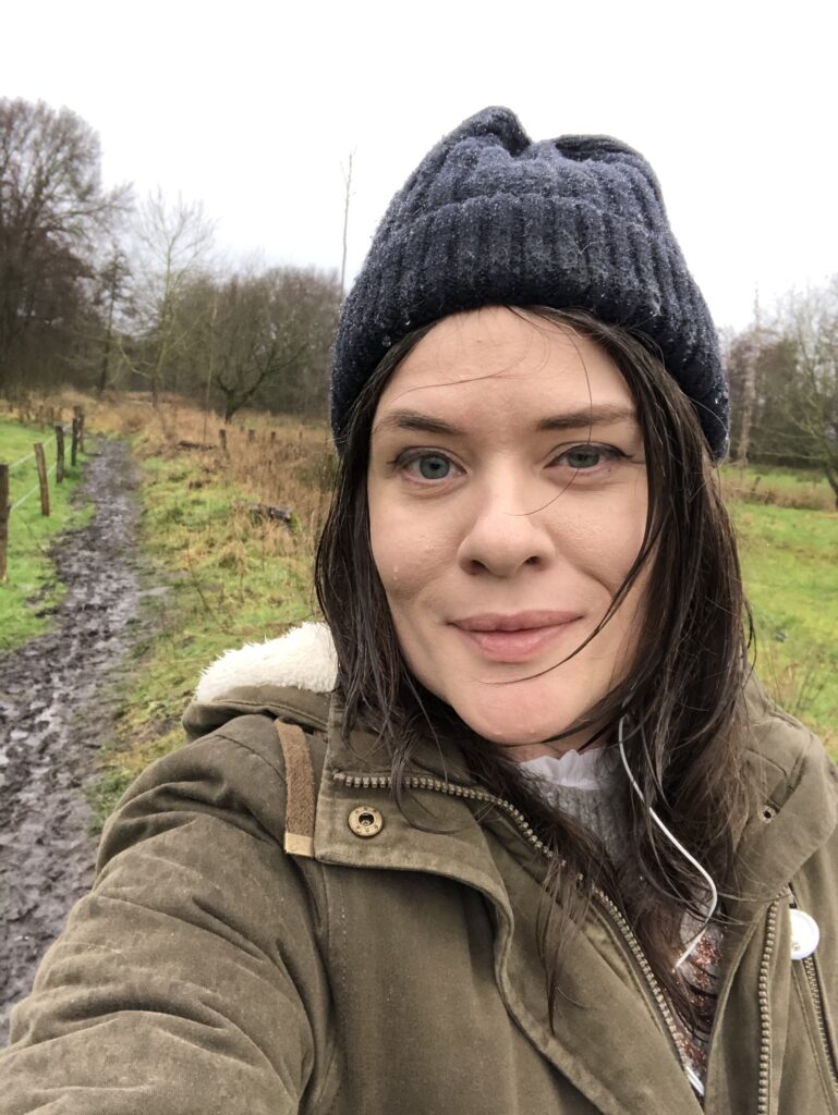 A woman walks a muddy path in a green coat and black beanie.  She is soaking wet
