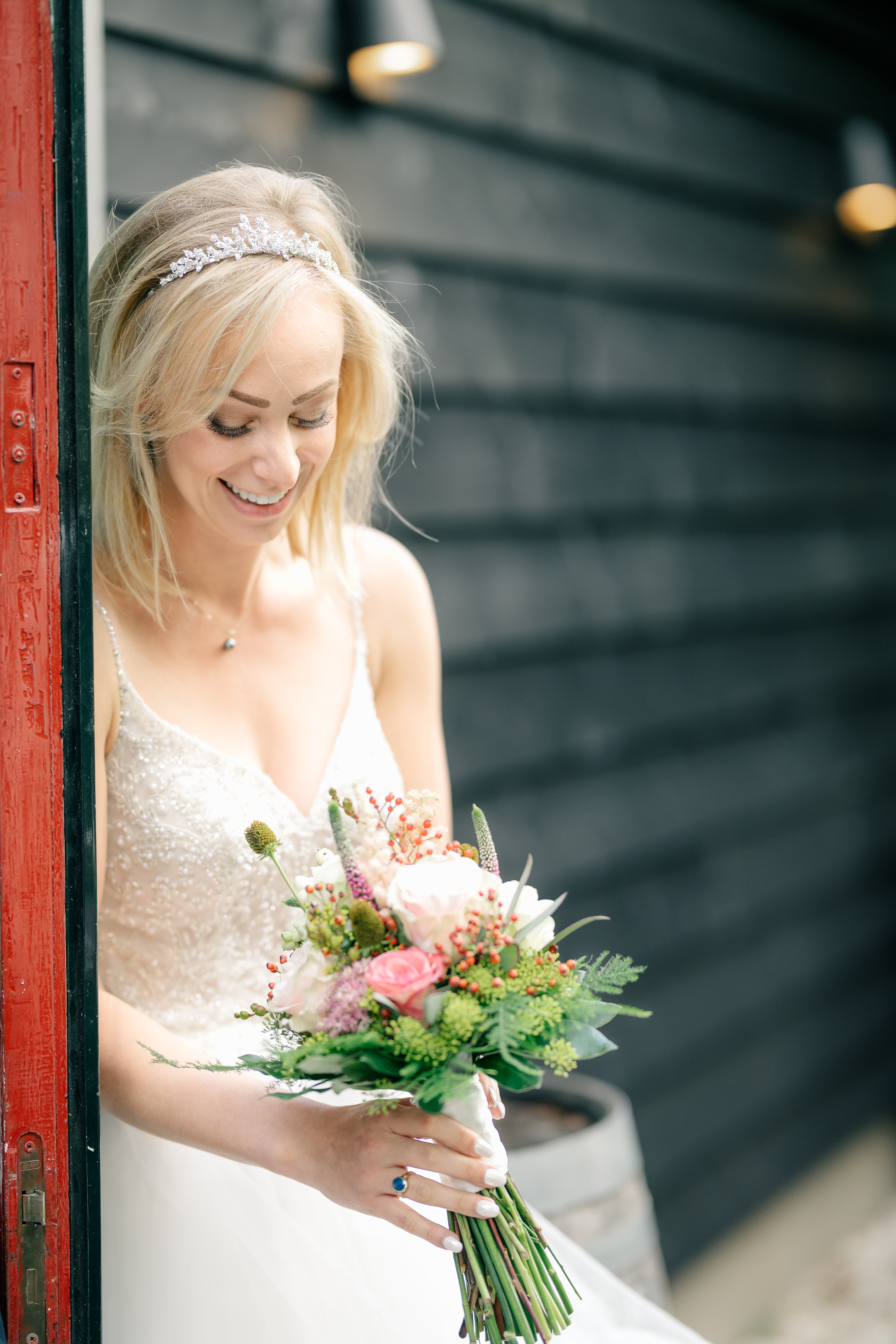The bride smiles and looks down at her bouquet standing on one side of a door in the First Touch before the ceremony