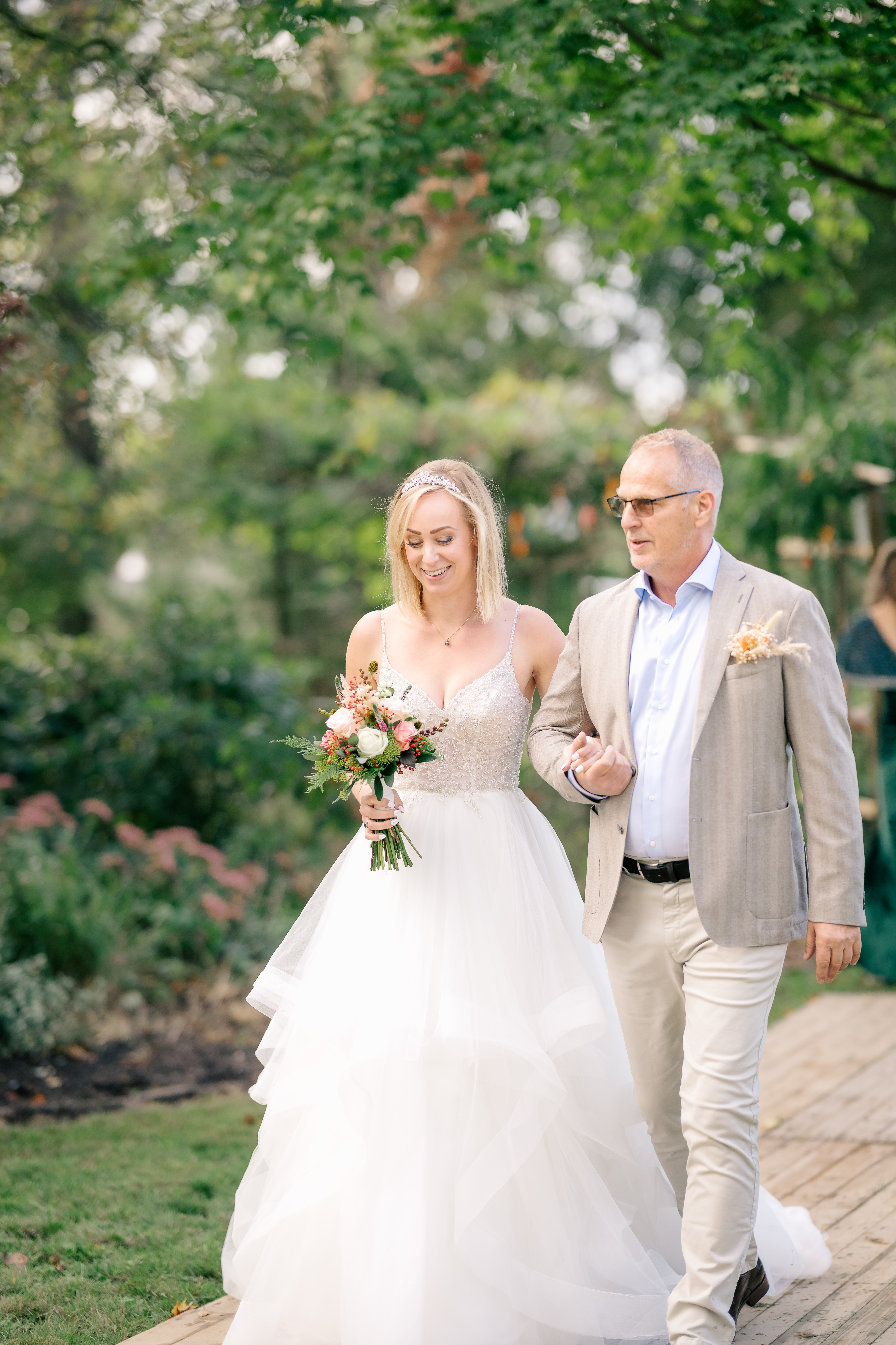 Father of the bride leads the bride down a wooden plank aisle in a september garden wedding in Heerle.  All the garden is still green