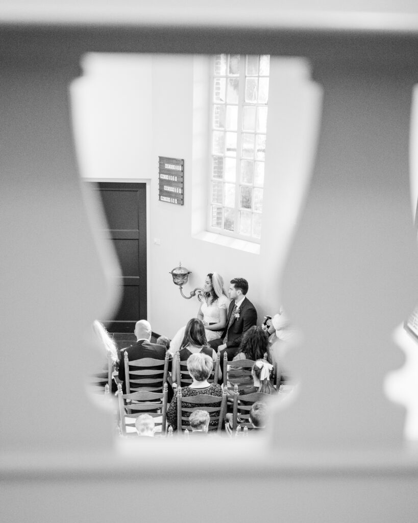 Black and white photo of wedding couple sitting during their ceremony taken through the balustrades of the choir seats