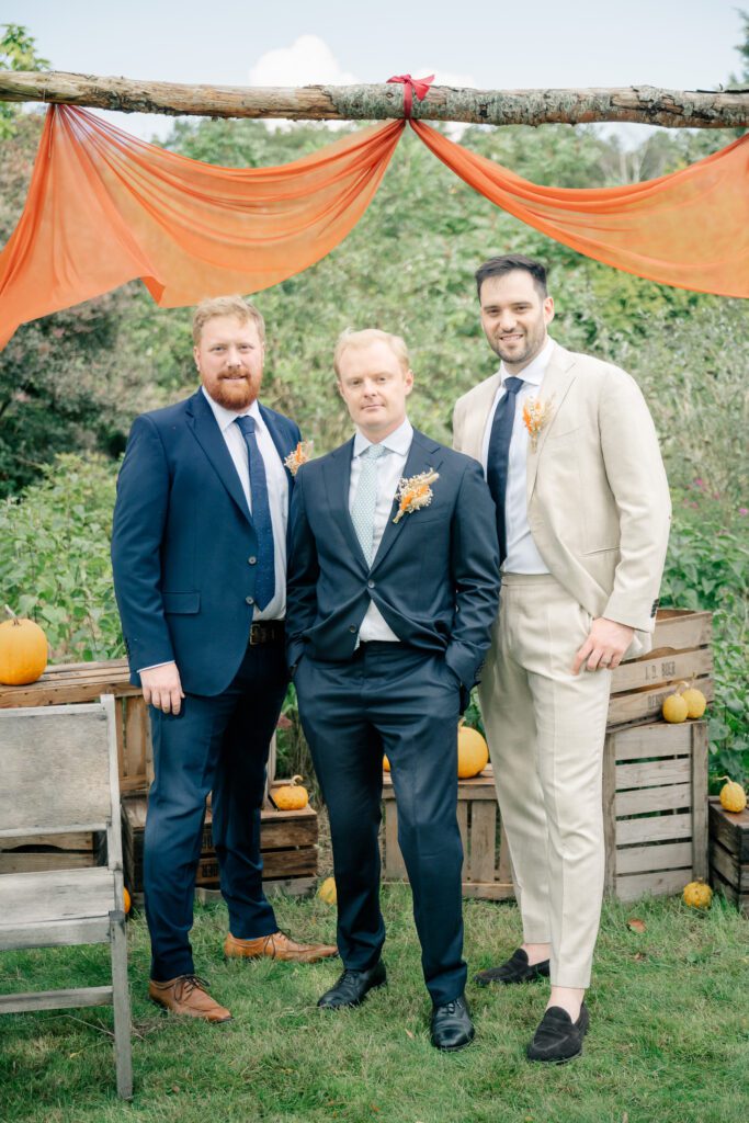 A groom stands under an orange bedecked arch flanked by his best men in a garden wedding