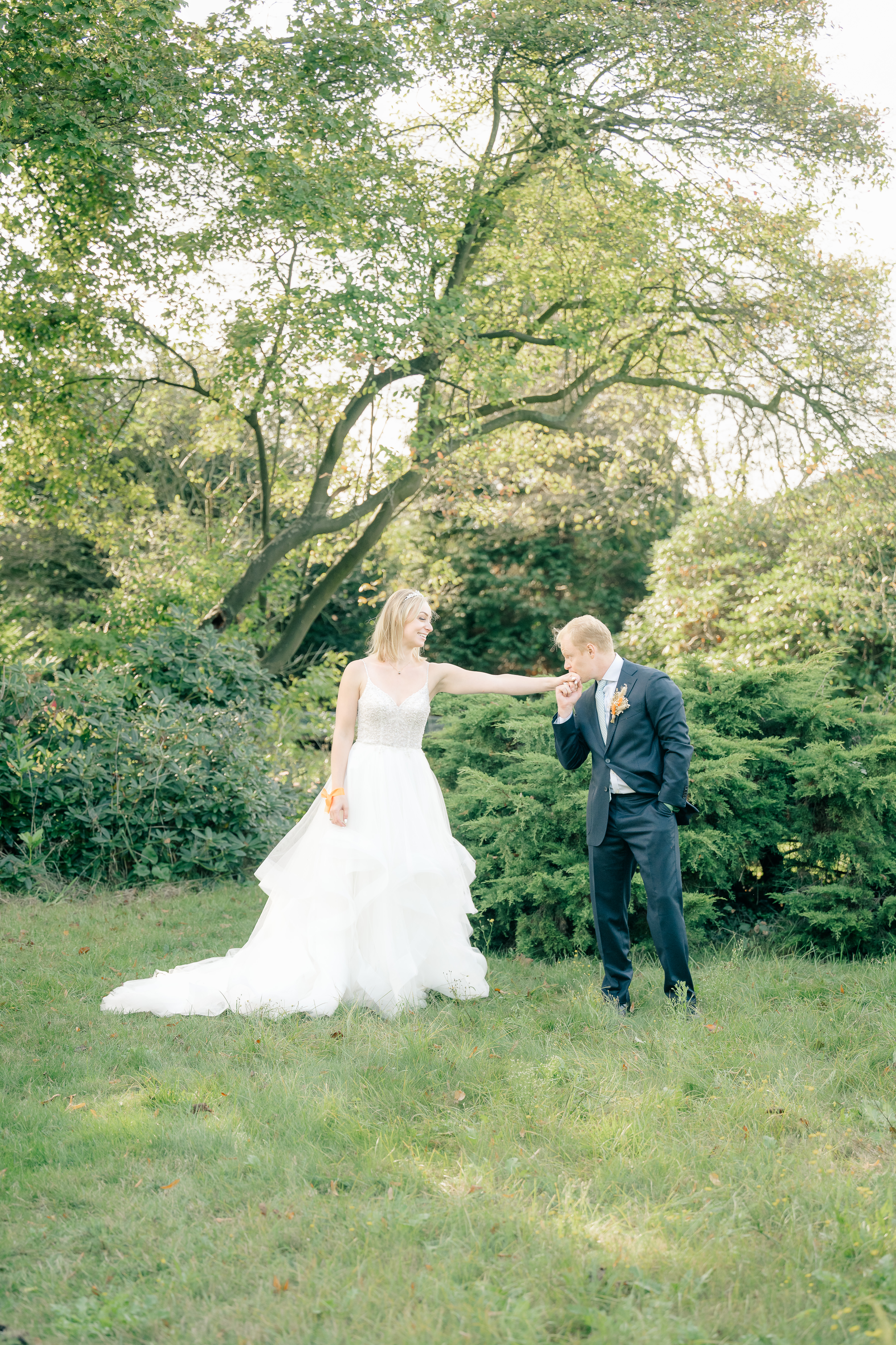 A bride and groom stand arm distance apart in a garden and the groom lifts the bride's hand to kiss it