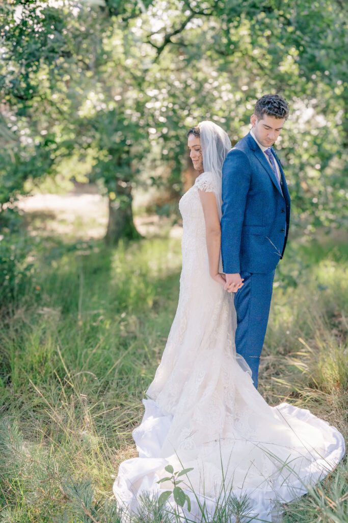 A bride and groom stand back to back holding hands in a shady nature area