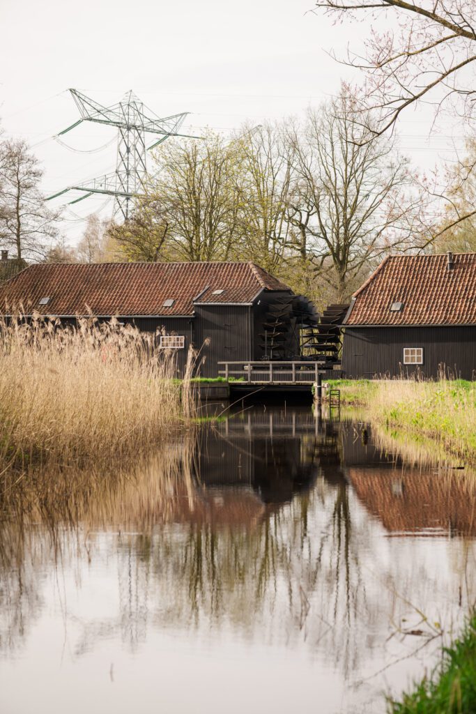 view of a black watermill by a pond with a red tile roof and reeds in front. the watermill reflects perfectly in the still water of the pond