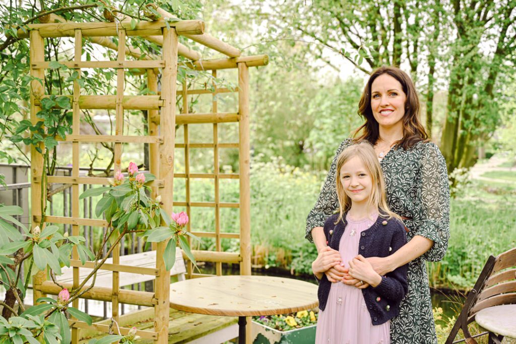 a mother and daughter stand posing for a photo in a lush green garden by a canal
