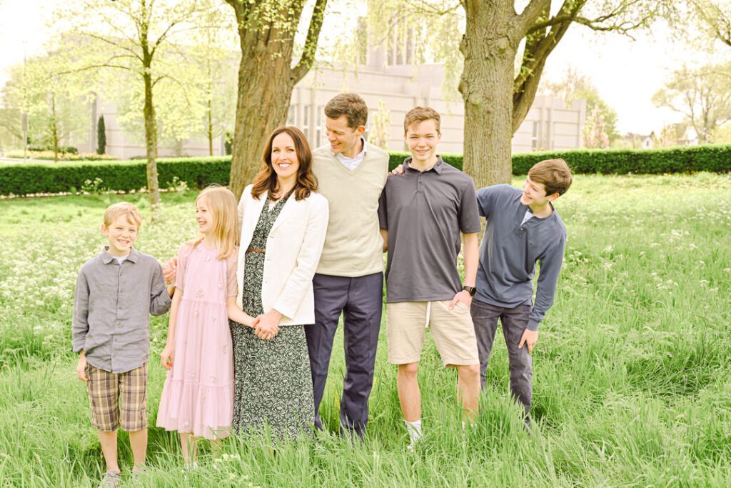 a family stands in long grass under trees with their leaves just budding, in front of a temple for the Church of Jesus Christ of Latter-Day Saints